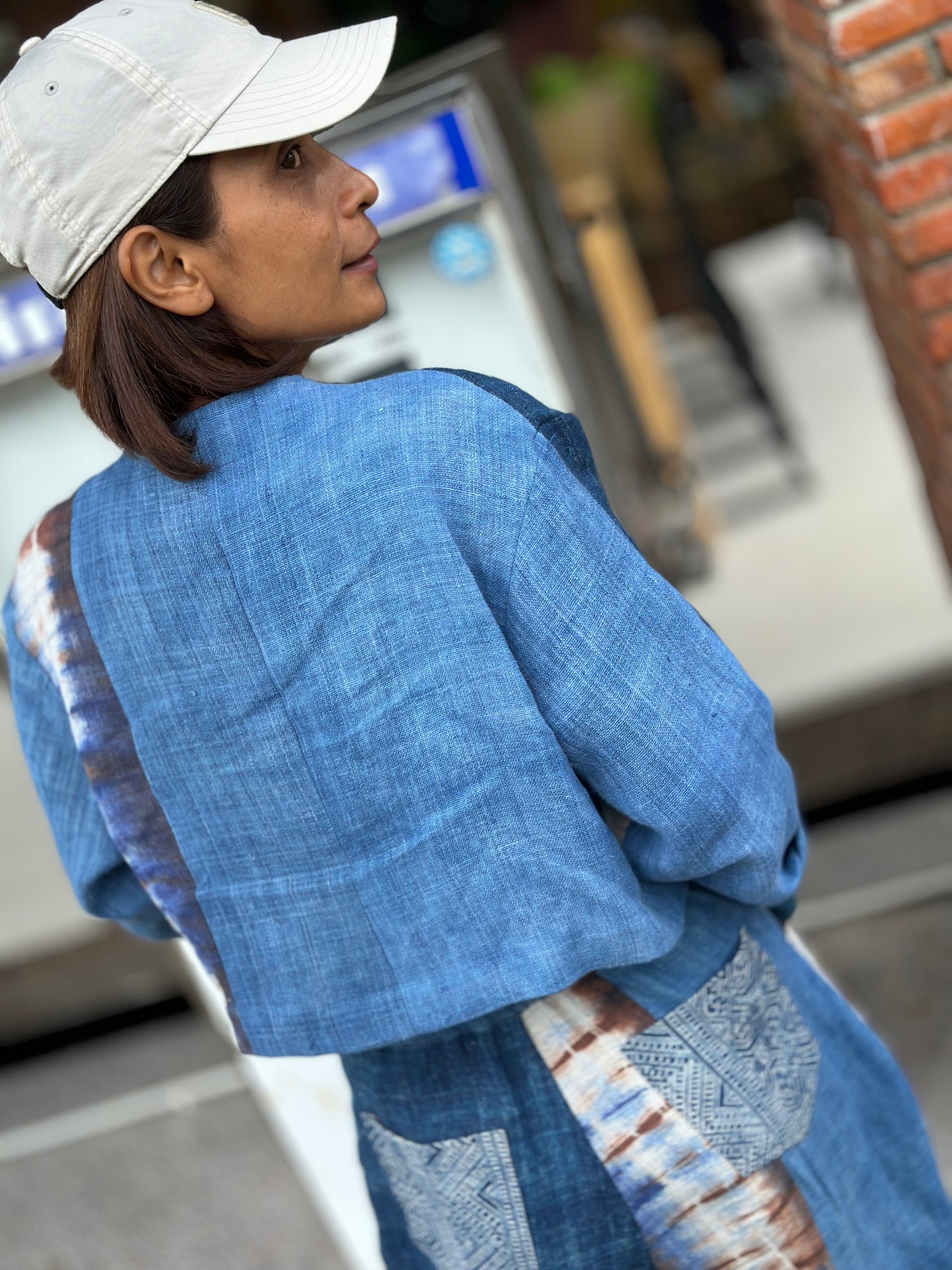 Nuichan patchwork jacket and pants set on a woman, featuring a indigo design with shibori and batik details, against a urban parking garage backdrop, with baseball cap and smartwatch. Back view