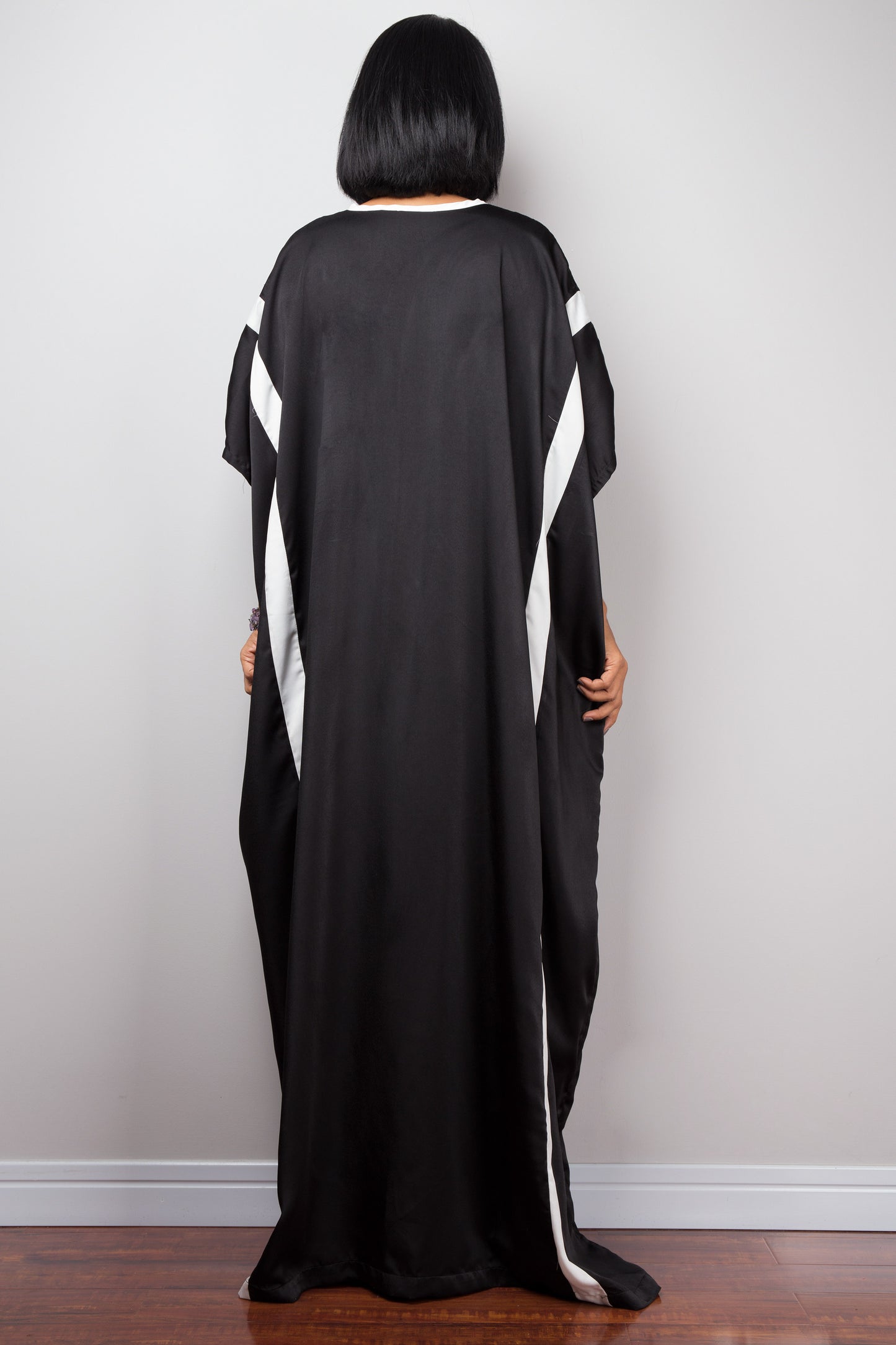 Nuichan’s black kaftan maxi dress with white stripe pattern, worn by a woman with dark hair, posed backwards against a gray wall on a wooden floor.