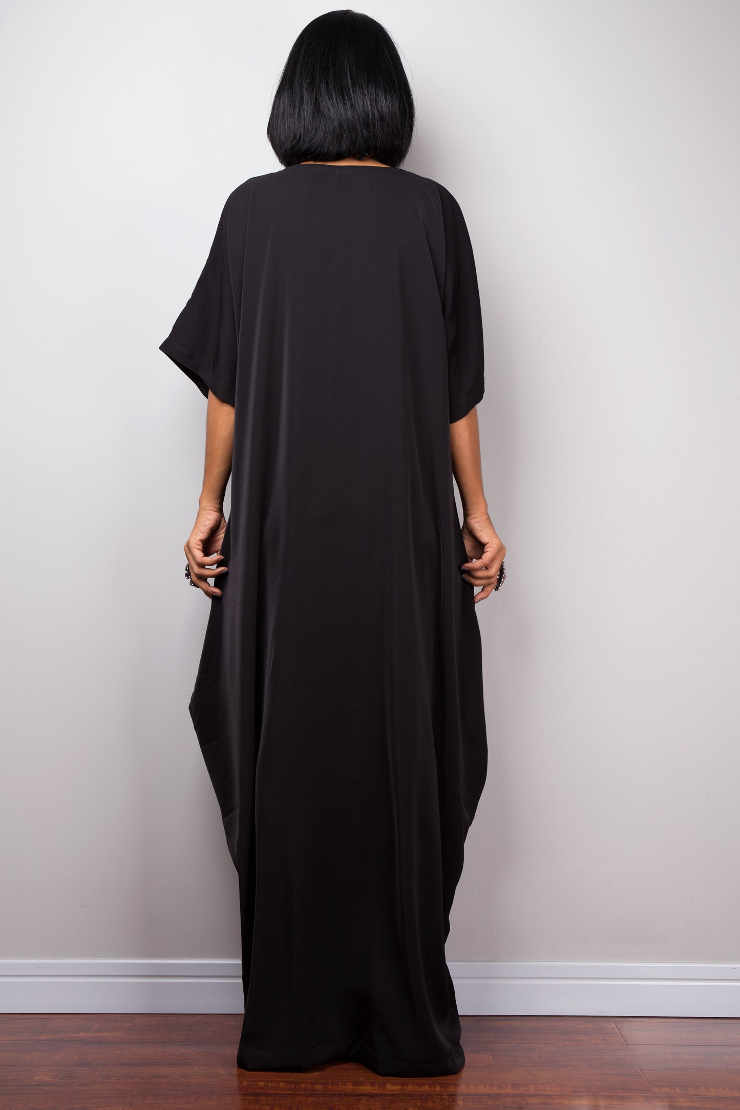 Black kaftan maxi dress by Nuichan, worn by a woman with short black hair, standing gracefully against a gray wall and wooden floor, back view.