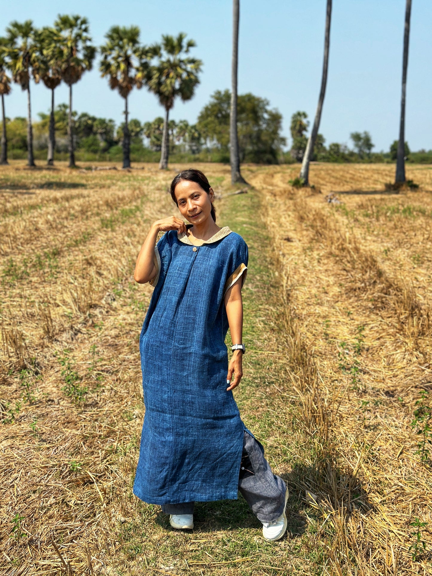 A woman modeling a blue hemp kaftan dress from Nuichan, with a loose fit, Peter Pan collar, and short sleeves, standing in a natural field setting with palm trees.