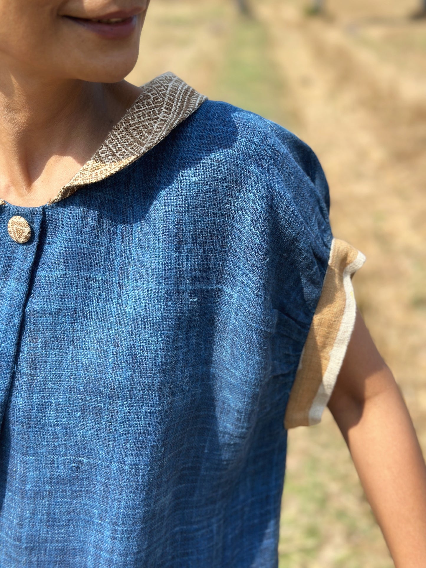 Nuichan’s blue hemp kaftan dress with brown earth-tone batik designs, worn by a woman with dark hair, posed in a scenic field with palm trees and dry grass, highlighting its boho style and Peter Pan collar.