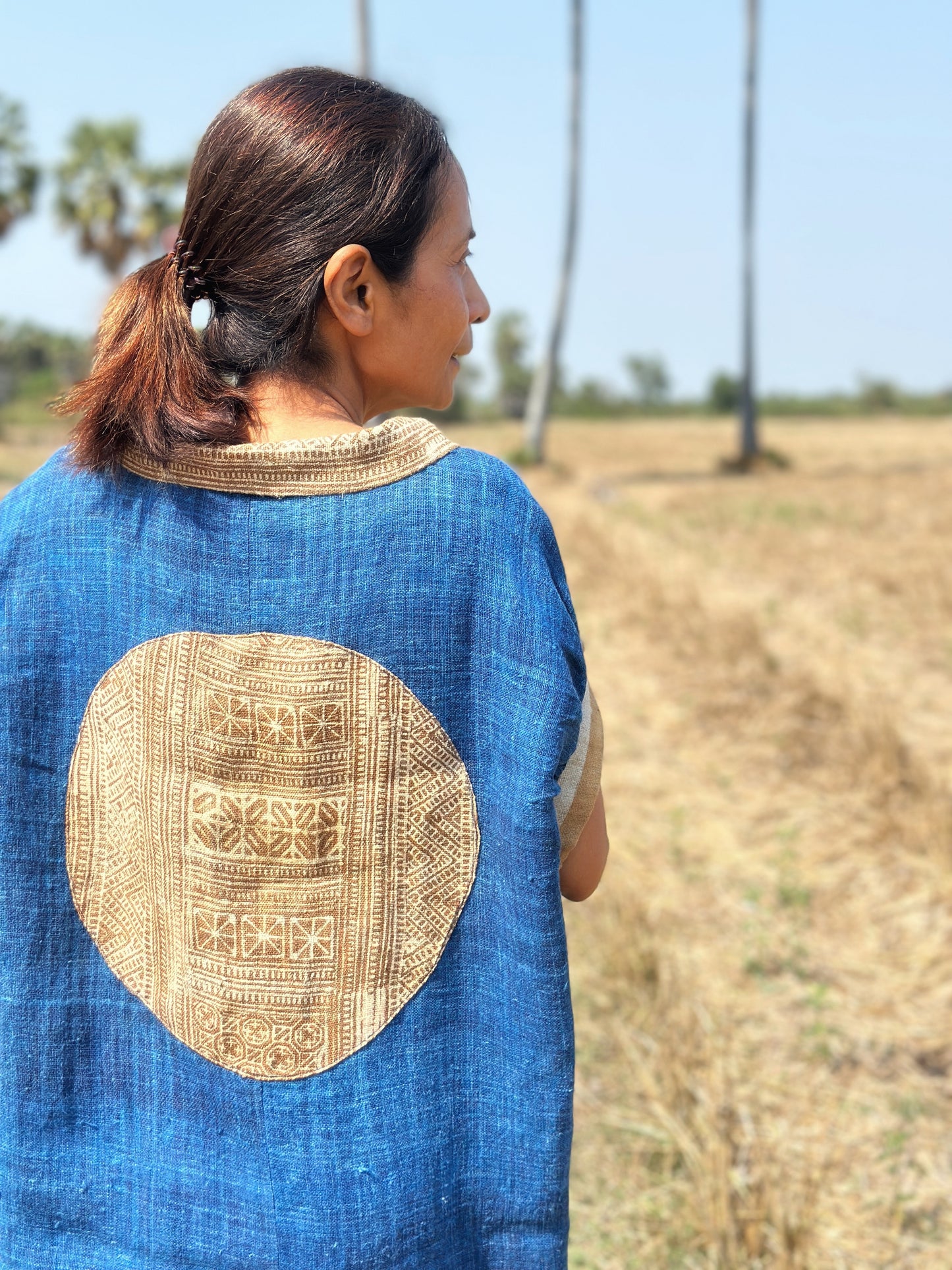 Blue hemp kaftan dress from Nuichan, worn by a woman with dark hair, posed in a dry field with palm trees, featuring its boho-chic design, flowing silhouette, brown earth-tone batik circle on the back, and Peter Pan collar.