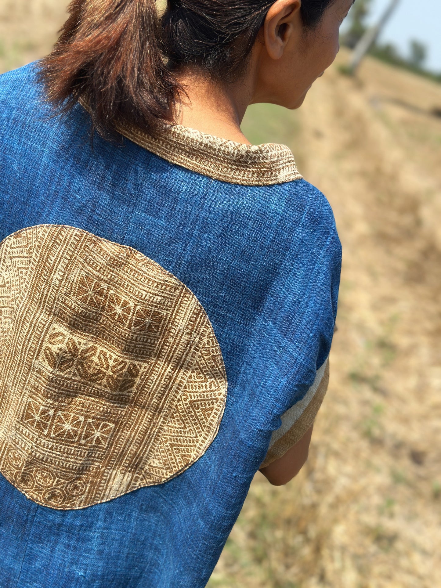 A floor-length blue hemp kaftan dress from Nuichan, worn by a woman from behind, featuring a large brown earth-tone batik circle on the back, set in a dry field with palm trees and blue sky, with a Peter Pan collar.
