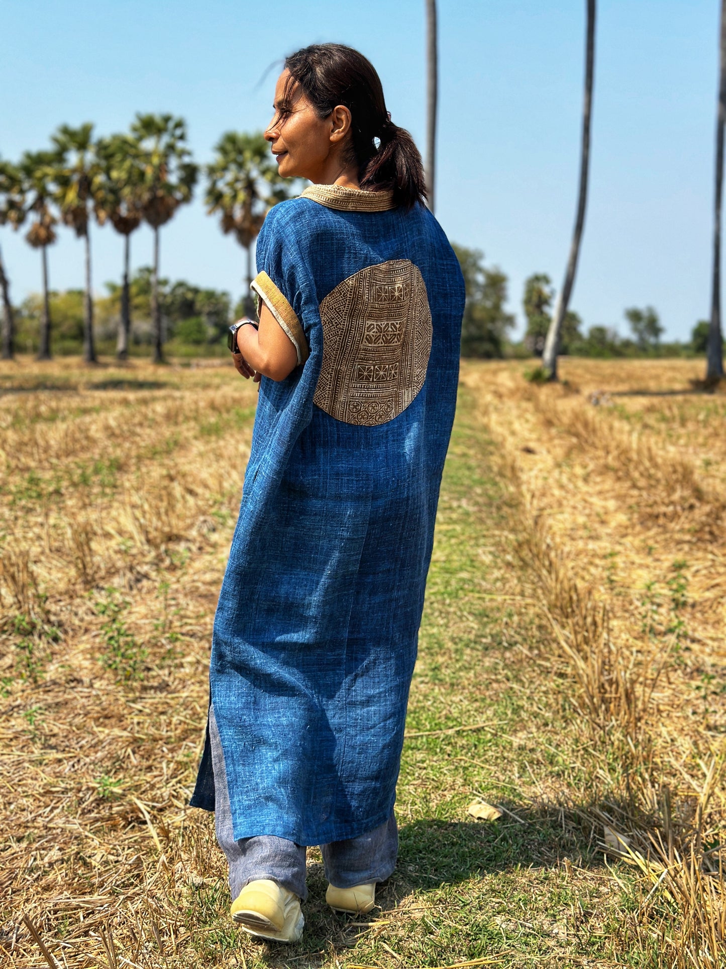 A woman modeling a blue hemp kaftan dress from Nuichan, with a loose fit, Peter Pan collar, and short sleeves, standing in a natural field setting with palm trees, featuring a brown batik circle on the back.
