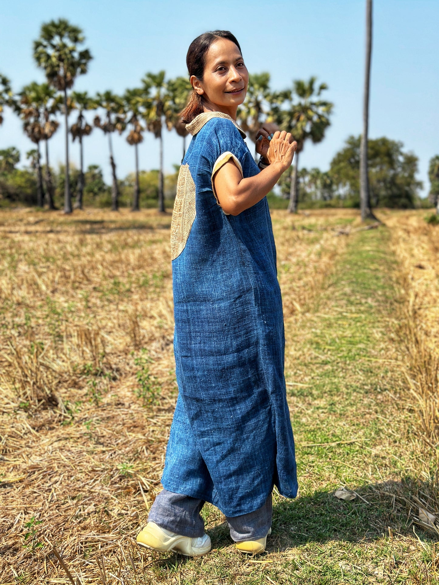 A woman wearing Nuichan’s blue hemp kaftan dress with brown batik details, standing in a scenic field, showcasing the dress’s loose fit, short sleeves, and Peter Pan collar, paired with white sneakers and a brown batik circle on the back.