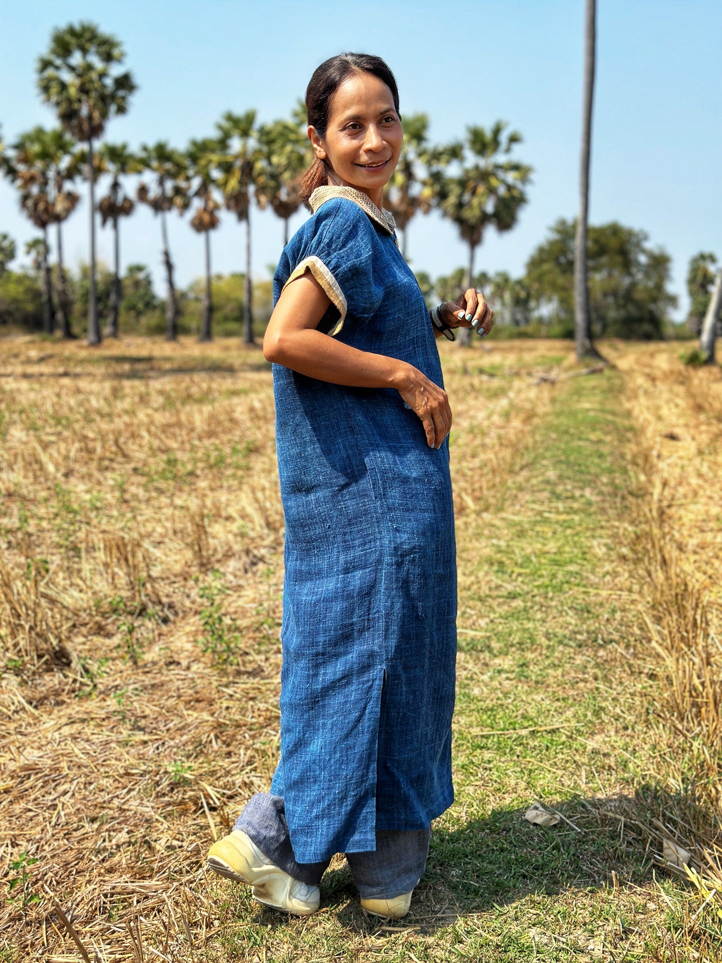 Nuichan’s floor-length hemp kaftan dress in blue with brown earth-tone batik motifs, modeled by a woman in an outdoor field with palm trees and dry grass, wearing a smartwatch, white sneakers, and a Peter Pan collar.