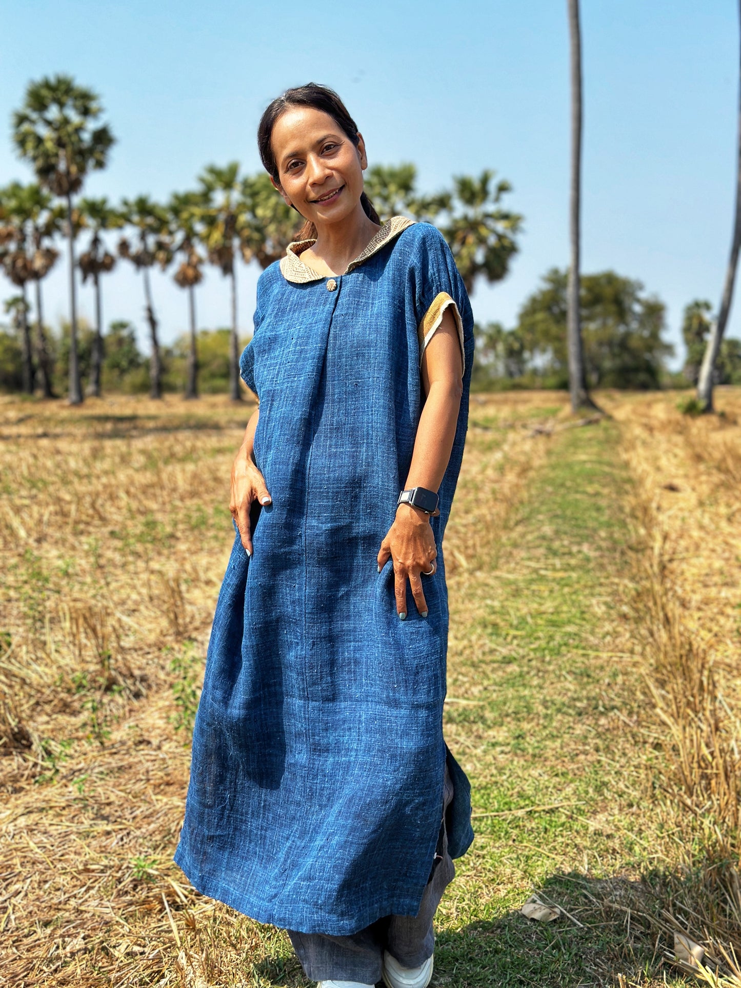 Woman in a blue hemp kaftan dress by Nuichan, standing in a field, showcasing the dress’s flowing silhouette, short sleeves, and brown batik Peter Pan collar, with palm trees and dry grass in the background.