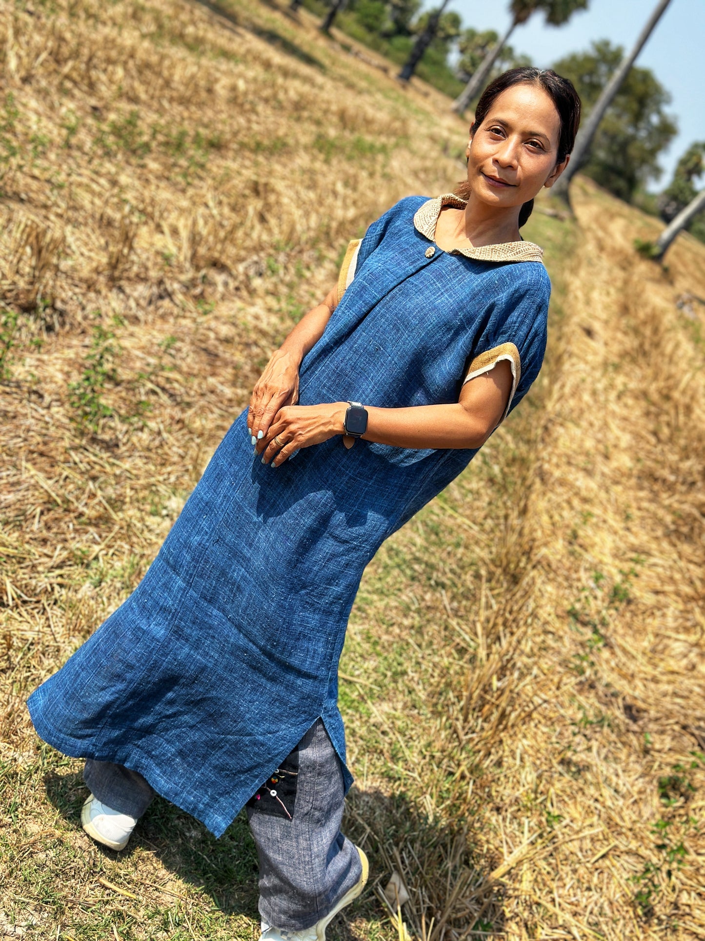 Blue hemp kaftan dress by Nuichan, worn by a woman with a ponytail, standing in a dry field with palm trees, featuring wide sleeves, a brown earth-tone batik circle on the back, and a Peter Pan collar.