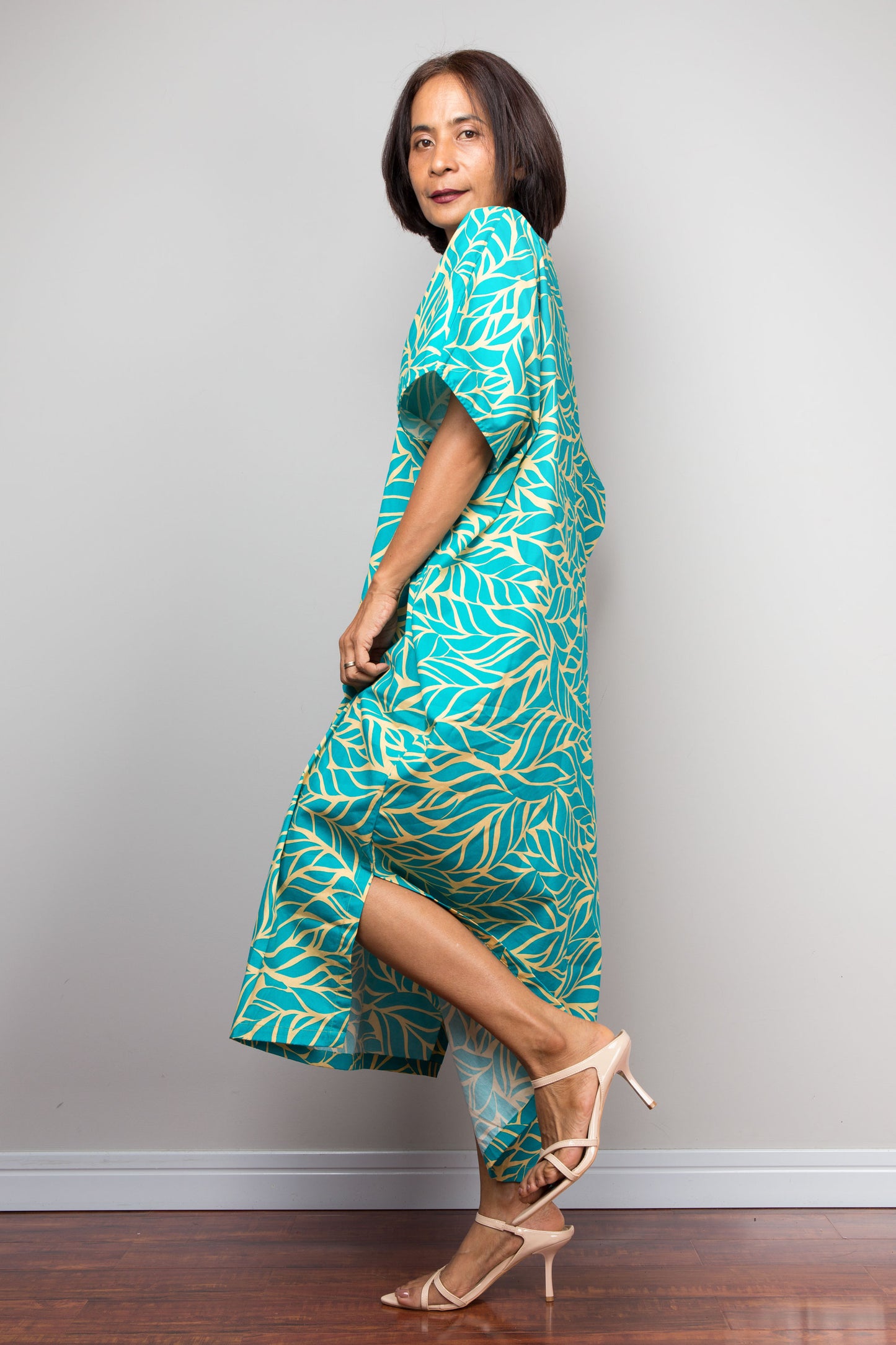 Petite Woman wearing an aqua green and yellow patterned kaftan dress against a gray wall.
