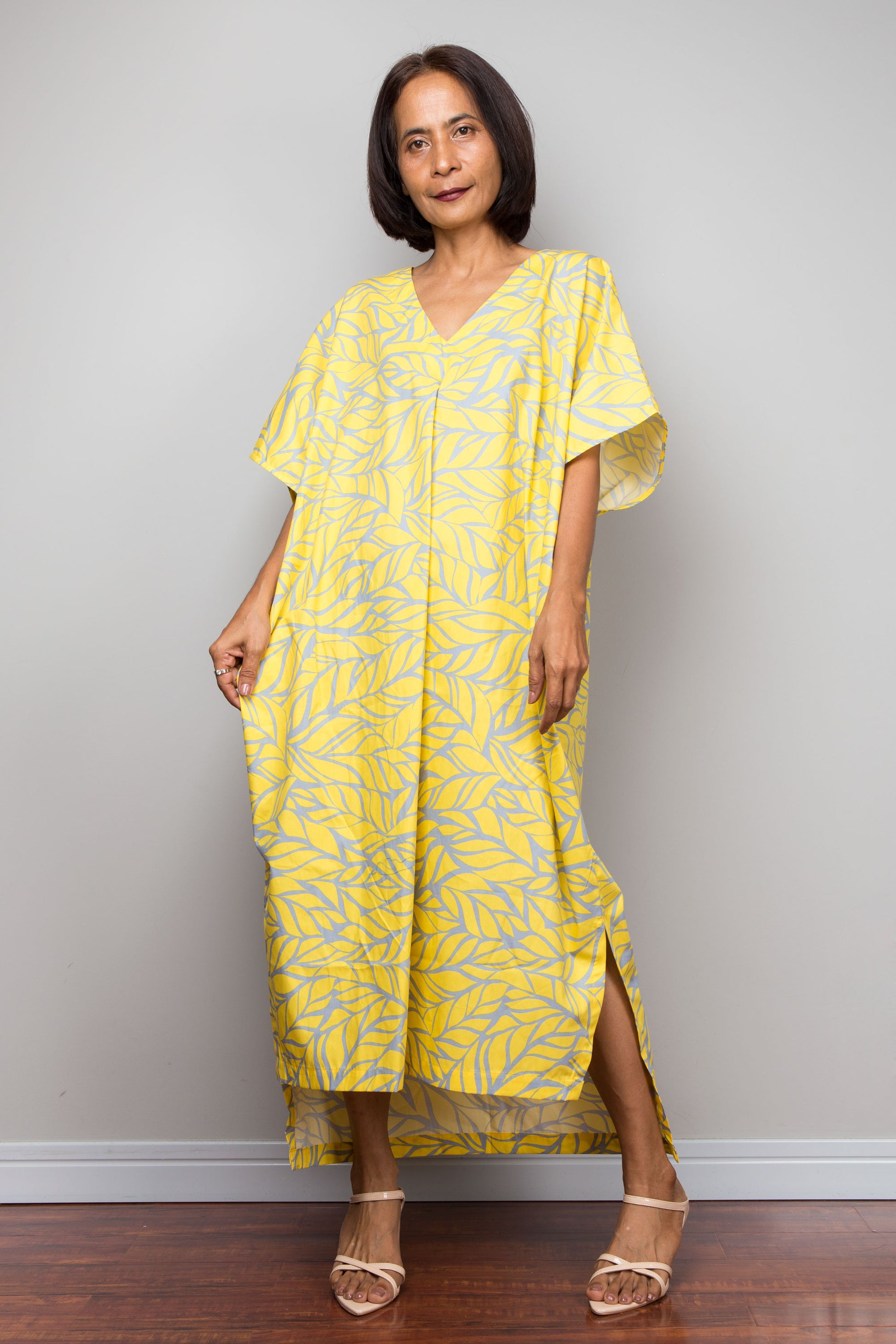 Woman wearing a yellow patterned, cotton caftan maxi dress standing against a plain wall.