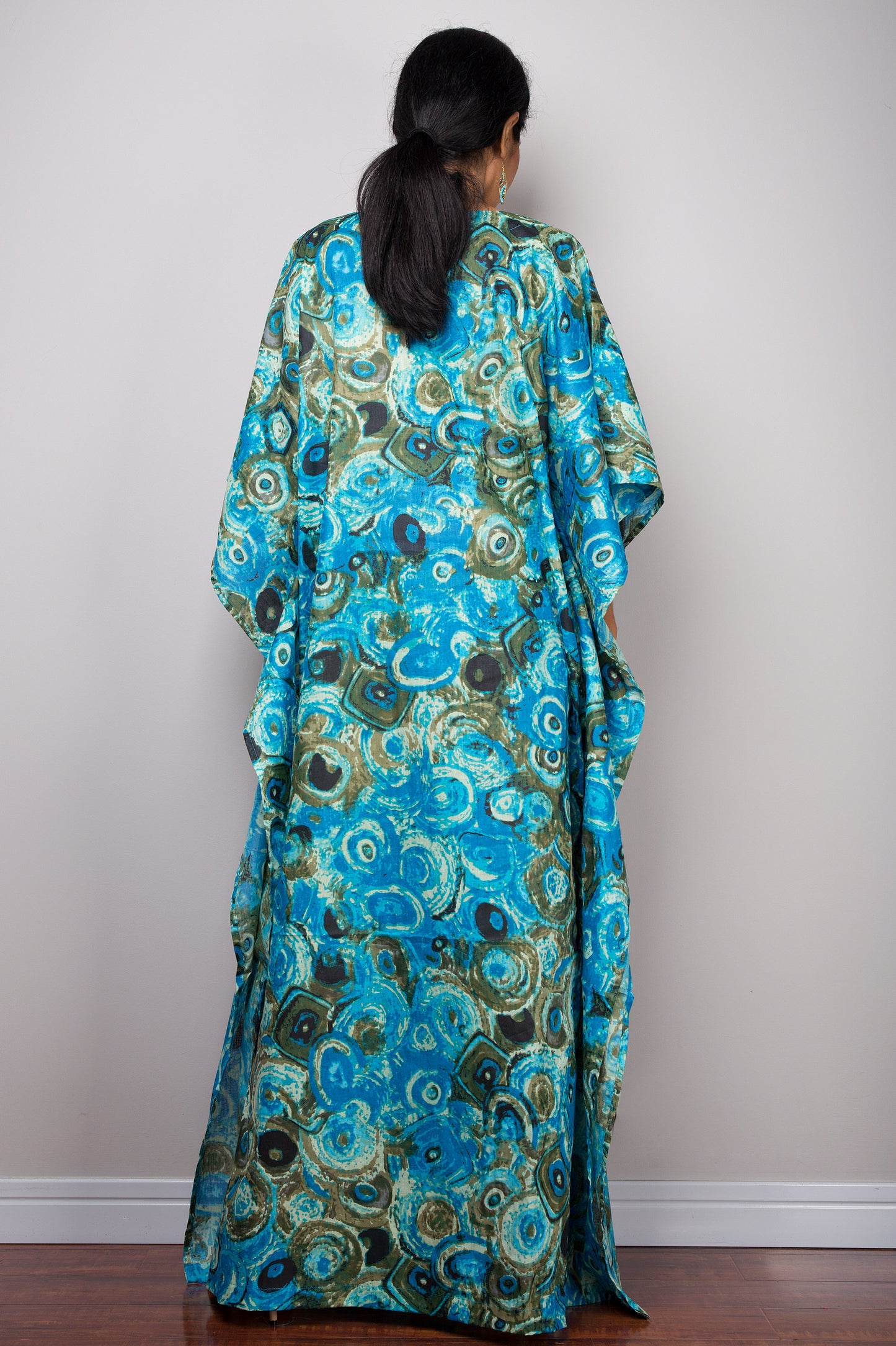 Model in a blue and green peacock pattern kaftan maxi dress featuring a deep V-neck and flared short sleeves, standing on a wooden floor with a gray wall background, accessorized with nude sandals and beaded bracelets.