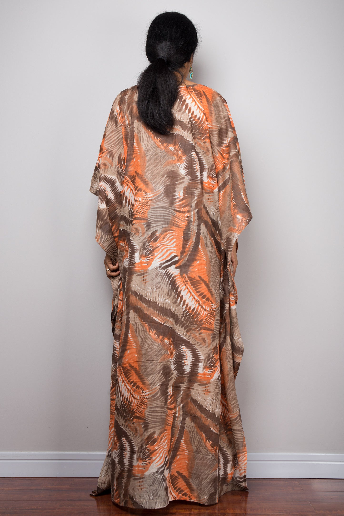 Model in an orange and brown abstract pattern kaftan maxi dress featuring a deep V-neck and flared short sleeves, standing on a wooden floor with a gray wall background, accessorized with nude sandals and beaded bracelets.