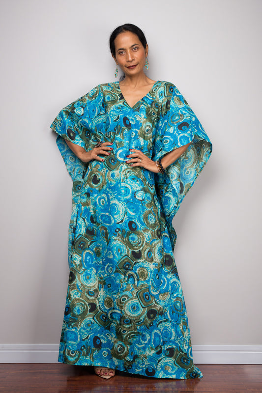 A woman wearing a blue peacock abstract kaftan maxi dress with a deep V-neckline and wide sleeves, standing against a gray wall with a wooden floor, accessorized with nude sandals, turquoise earrings, and beaded bracelets.