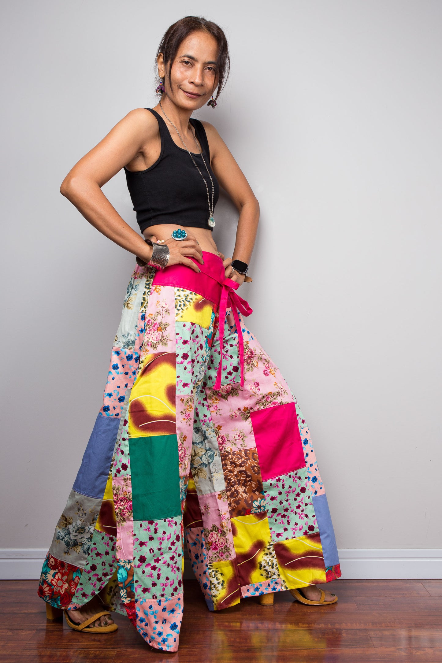 Three quarter side view of a petite model wearing the Nuichan pastel patchwork wide leg palazzo pants, showcasing the hot pink self-tie sash waistband, full ultra-wide leg silhouette, and hand-sewn quilt-style patchwork construction in mixed cotton satin pastel prints including mint, blush pink, teal, yellow satin and floral panels, styled with a black crop top