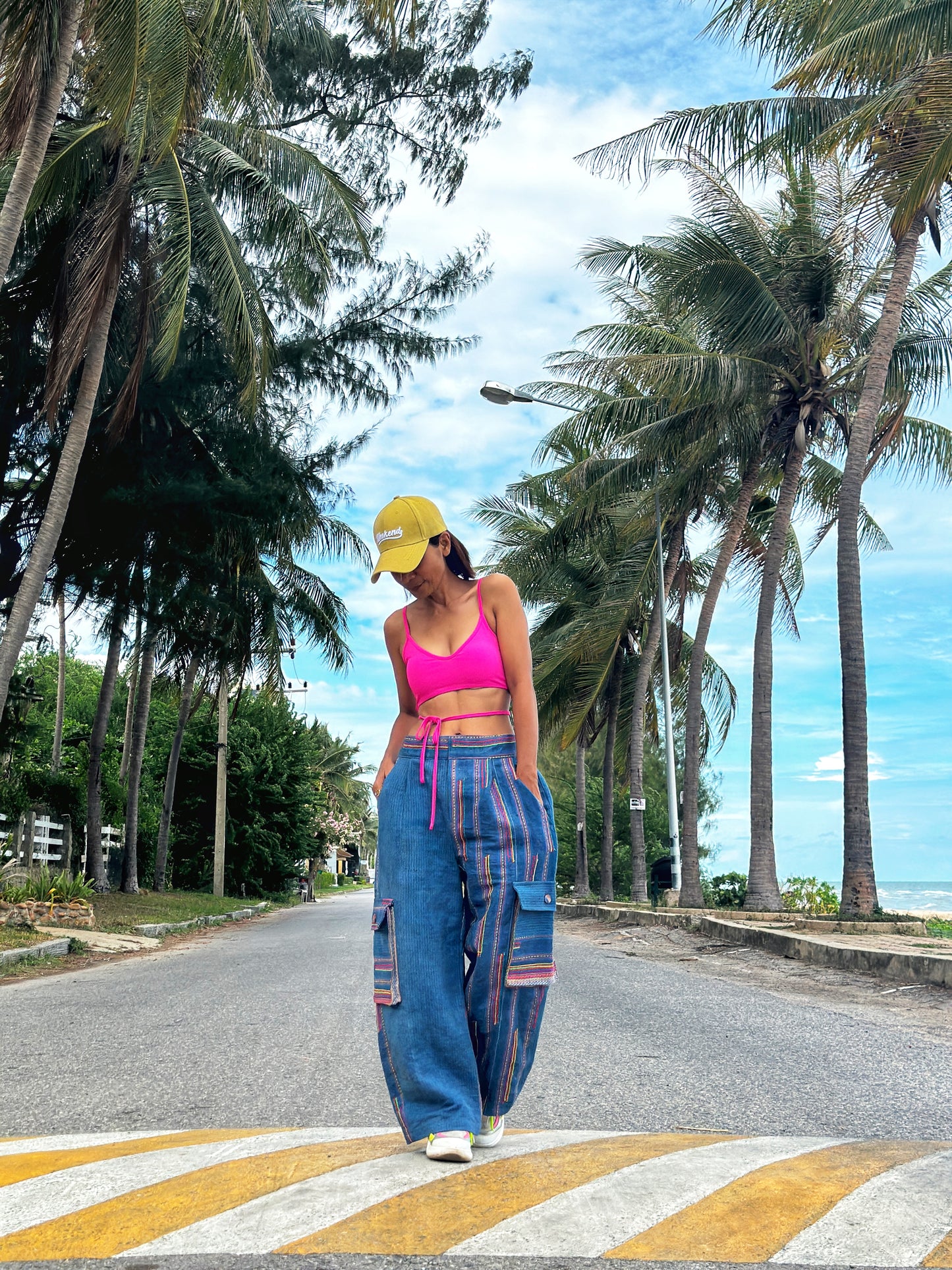 A person standing on a crosswalk wearing blue handwoven hemp cargo pants with a colorful top and a yellow hat, with palm trees and a clear blue sky in the background.