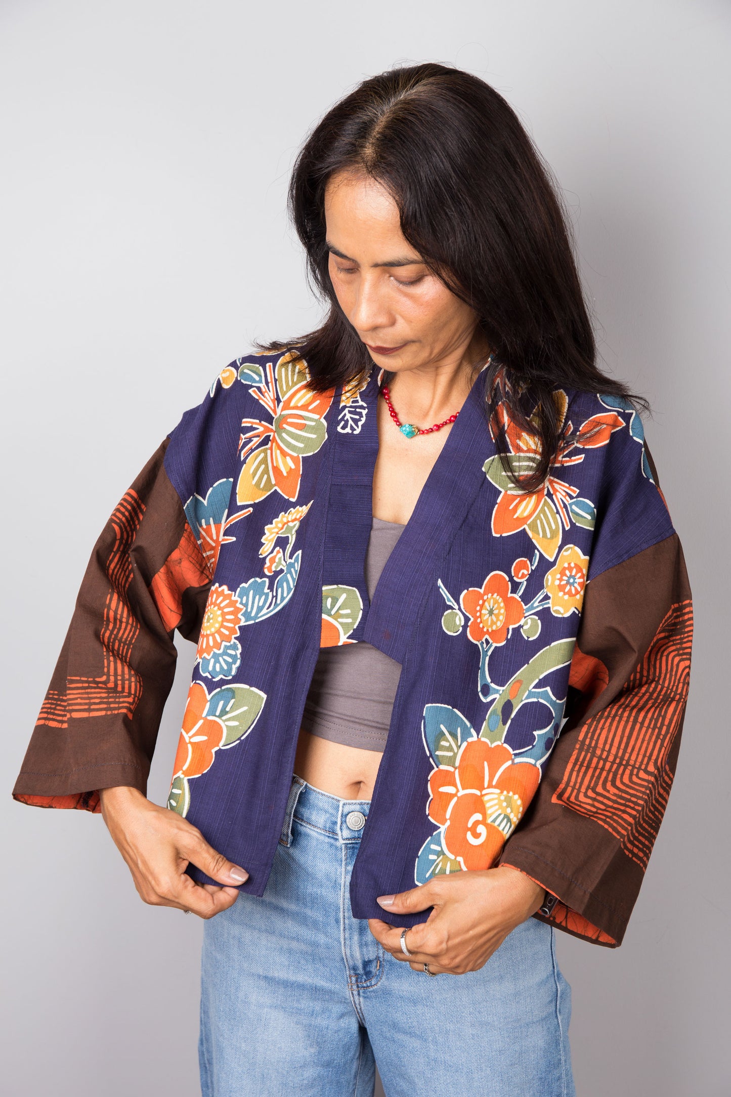 Woman wearing a colorful floral kimono jacket top over a gray top and blue jeans on a plain background