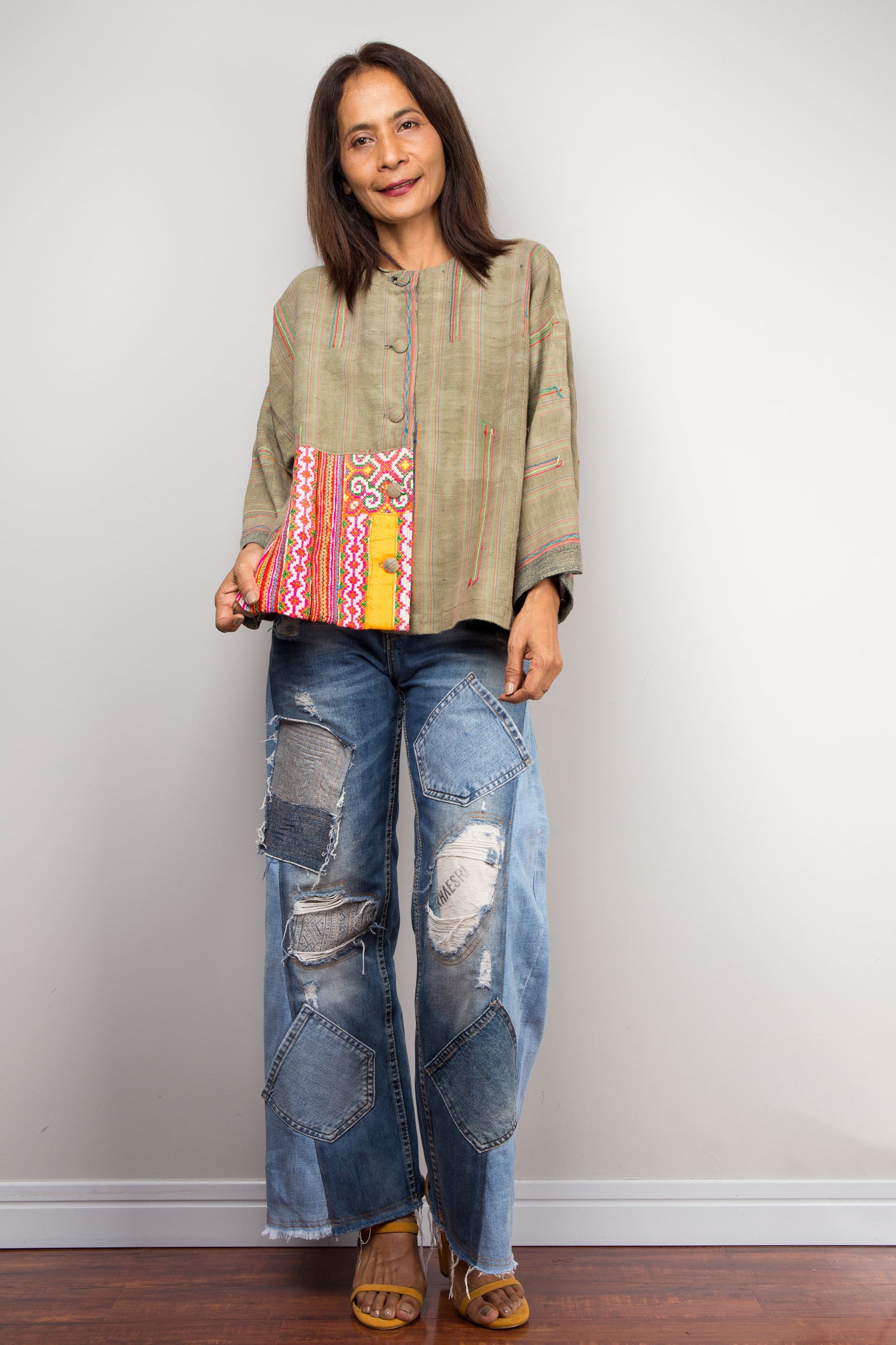Olive hemp jacket with flared sleeves and patchwork by Nuichan on a woman, paired with distressed jeans, set against a gray wall and wooden floor, with tan heeled sandals.