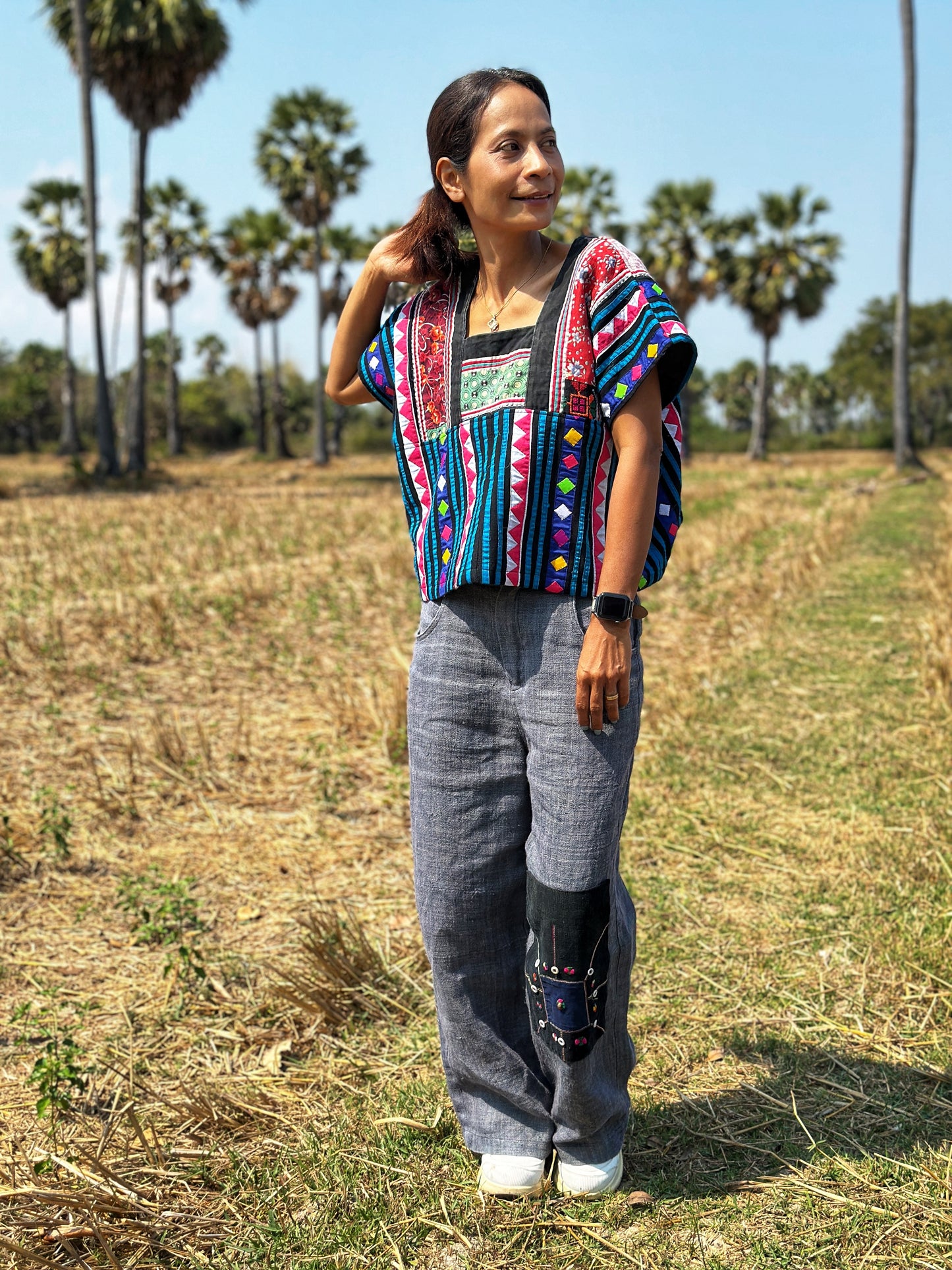 A cropped black, blue, magenta, and red Hmong tribal top from Nuichan, worn by a woman standing sideways, featuring loose, flowing fabric and short sleeves, set in a natural field.