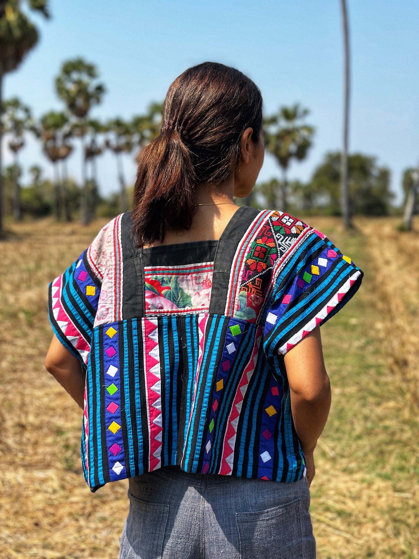 Black, blue, magenta, and red Hmong tribal crop top from Nuichan, worn by a woman with dark hair and a white smartwatch, back view posed in a natural field with grass and trees, featuring its boho-chic design and flowing silhouette.