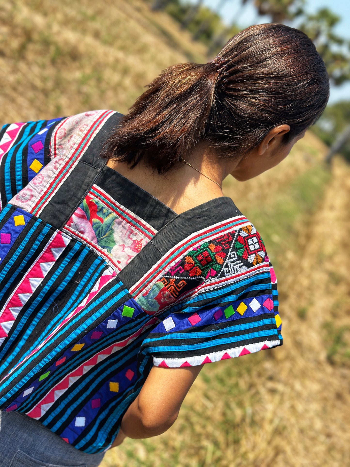 Nuichan’s cropped Hmong tribal top in black, blue, magenta, and red, modeled by a woman in an outdoor field with dry grass and palm trees. Back view.