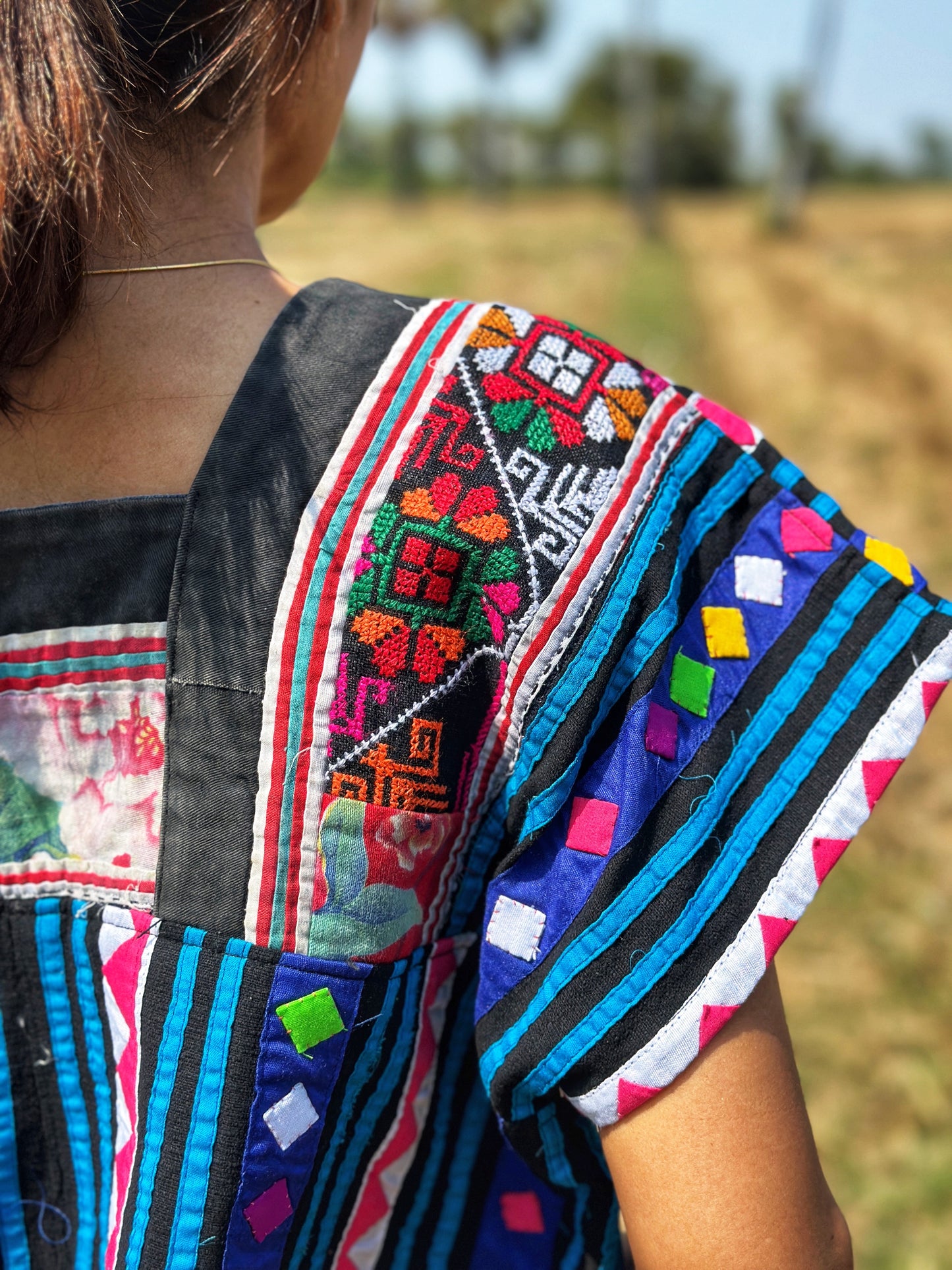 Nuichan’s cropped Hmong tribal top in black, blue, magenta, and red, modeled by a woman in an outdoor field with dry grass and palm trees. Close up view of the back shoulder part.
