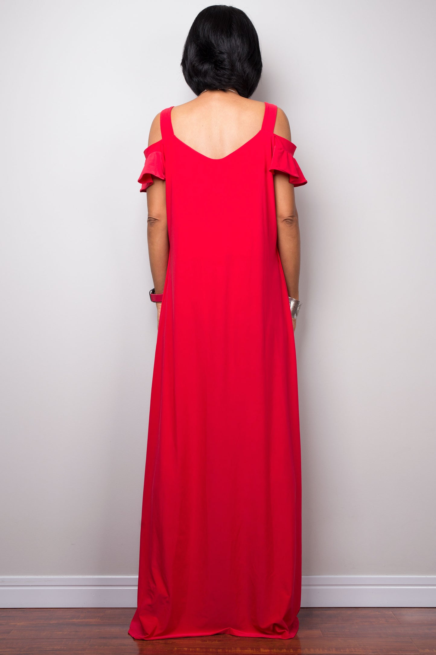 Woman showcasing a red cold-shoulder ruffle-sleeve kaftan maxi dress by Nuichan, standing on a wooden floor with a gray wall backdrop, accessorized with silver jewelry and nude sandals.