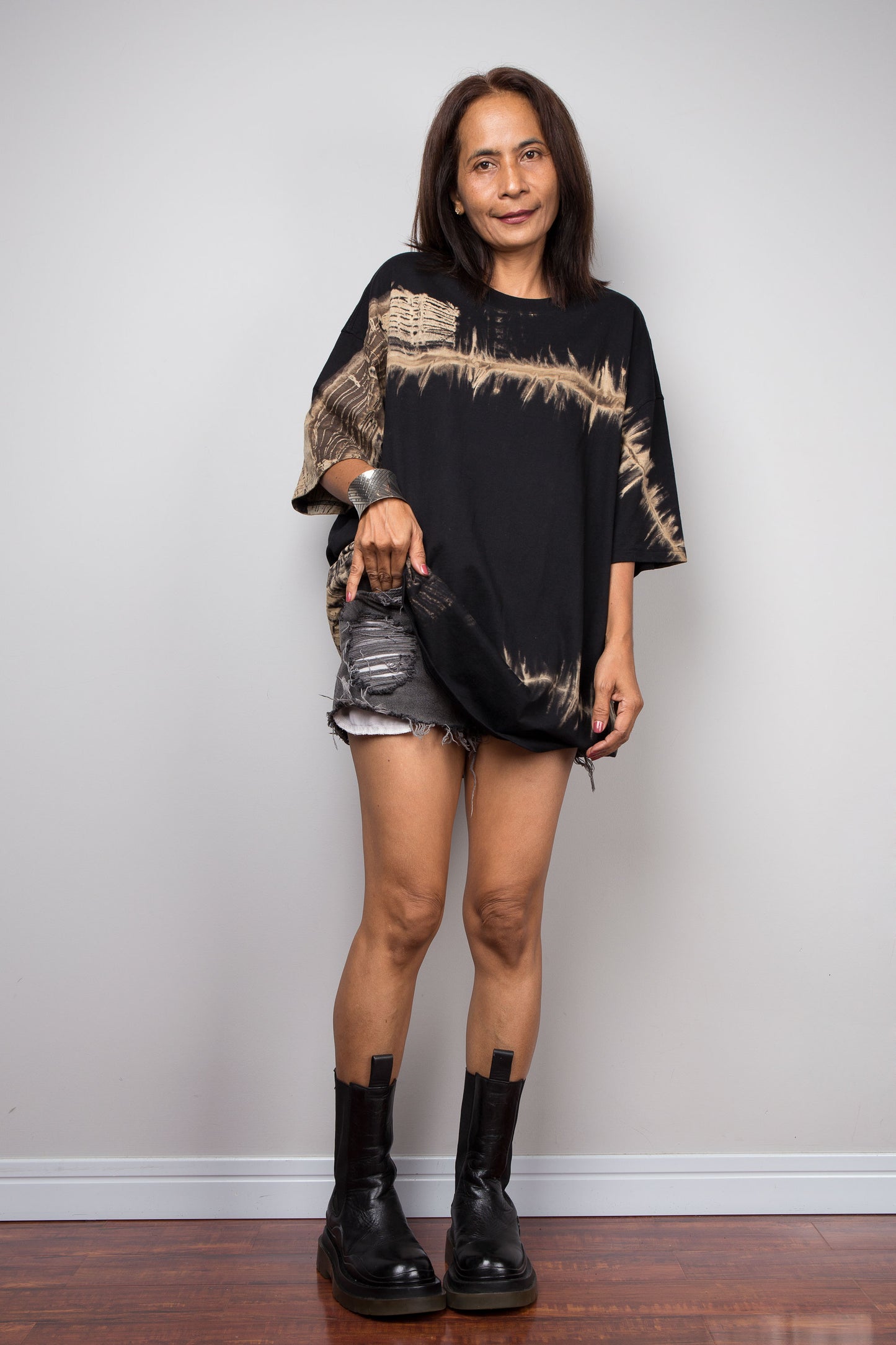 A woman modeling a black oversized short-sleeve t-shirt with a distressed beige graphic print across the chest, paired with distressed denim shorts and black leather boots, standing against a plain light gray wall.