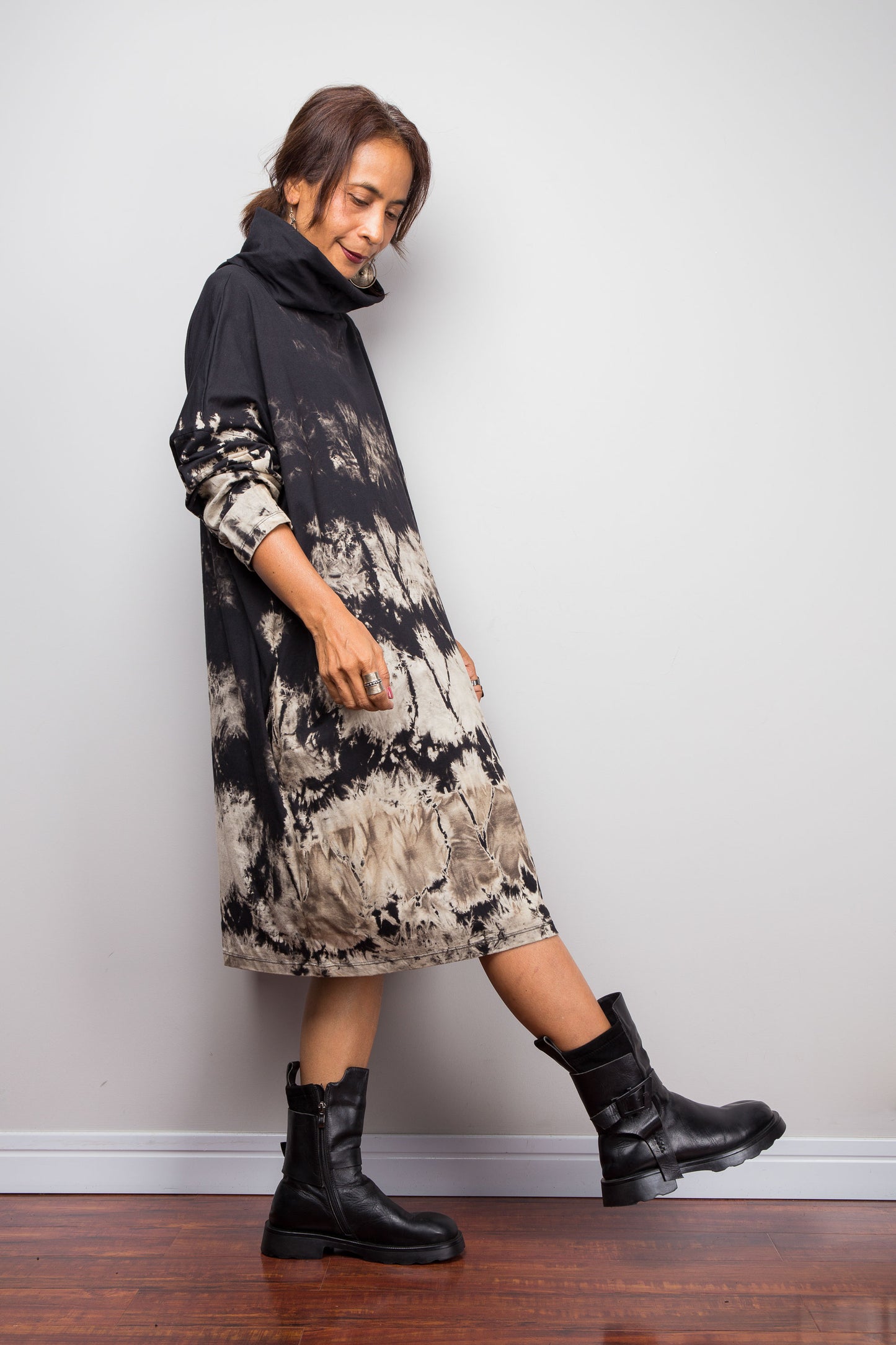 Full front view of a petite model wearing Nuichan's oversized reverse tie-dye tunic dress with a high funnel cowl neck and horizontal abstract sand-colored patterns.