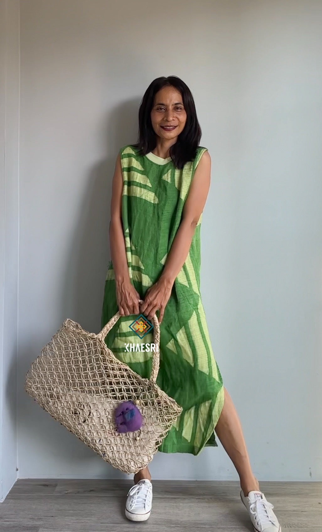 Close-up of a Nuichan sleeveless hemp kaftan dress, showcasing a handwoven green fabric with white geometric triangles. This artisanal boho dress, styled with a woven bag, reflects sustainable craftsmanship in a minimalist setting.