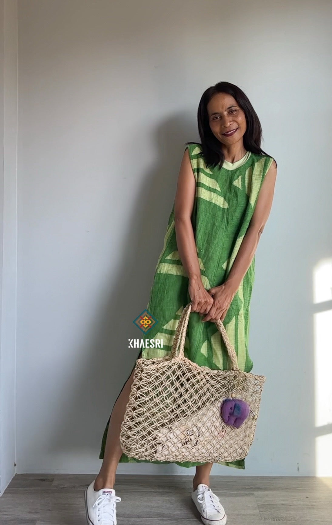 Woman styling a Nuichan sleeveless hemp kaftan dress with a green geometric pattern, paired with white sneakers and a woven bag. This handwoven boho dress, photographed indoors, offers a chic, sustainable look with practical side pockets