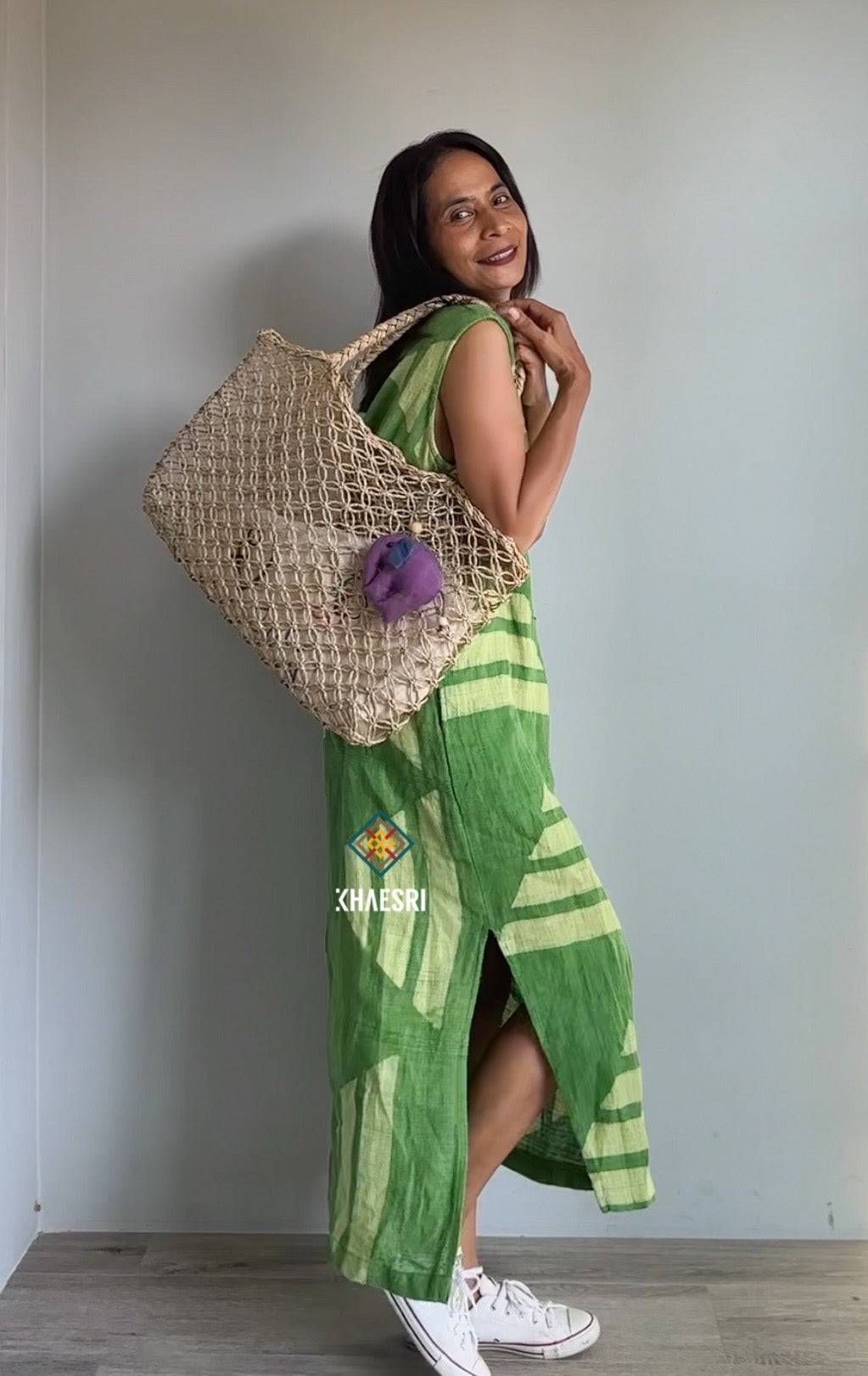 Woman wearing a sleeveless hemp kaftan dress by Nuichan, with a handwoven green geometric design. This boho dress, ideal for beach cover-ups or casual outings, is styled with white sneakers and a woven bag indoors.