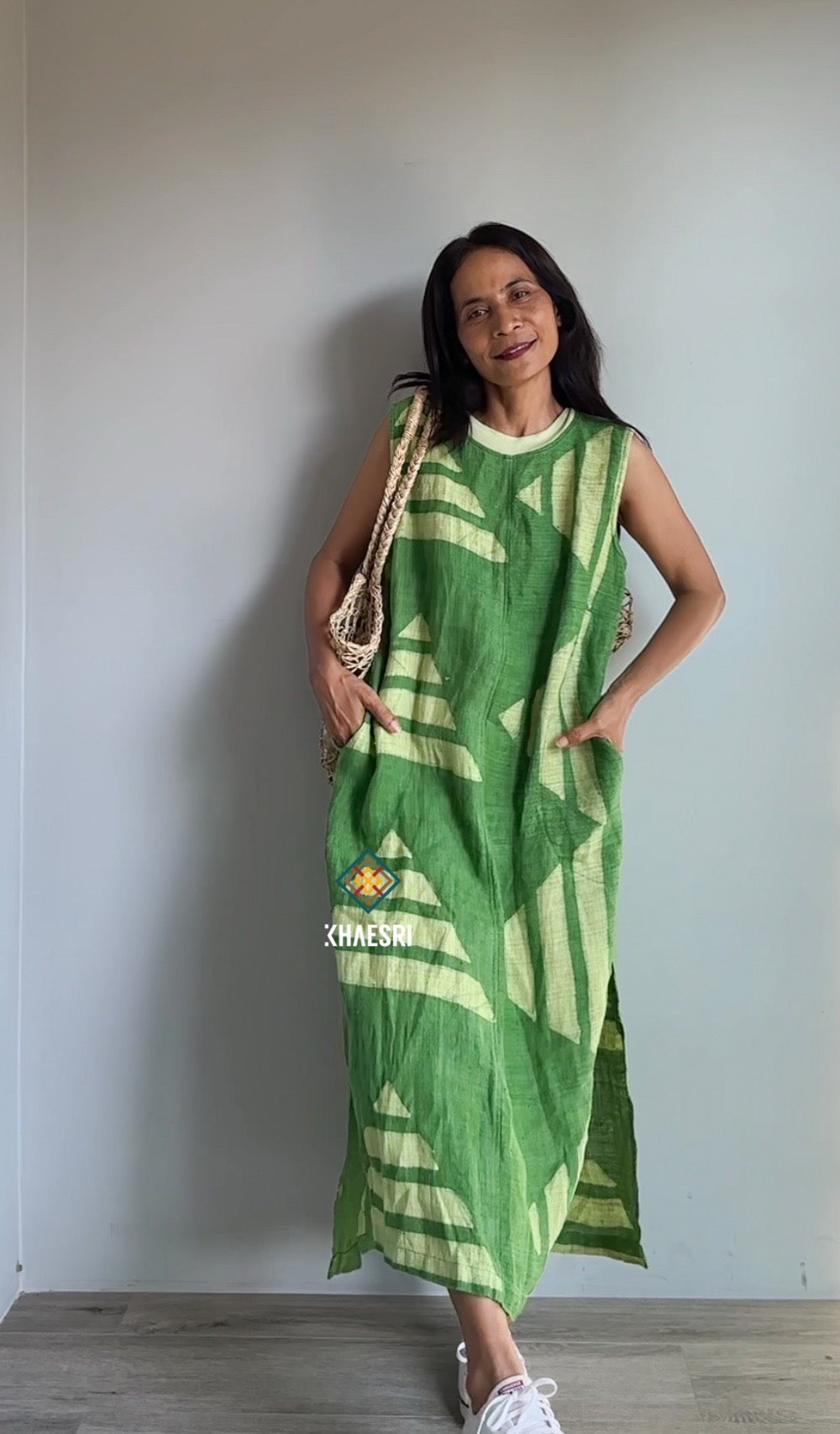 Model in a Nuichan sleeveless hemp kaftan dress, featuring a vibrant green fabric with white geometric patterns. This handwoven boho dress, captured indoors with a woven bag, exudes an earthy, artisanal aesthetic