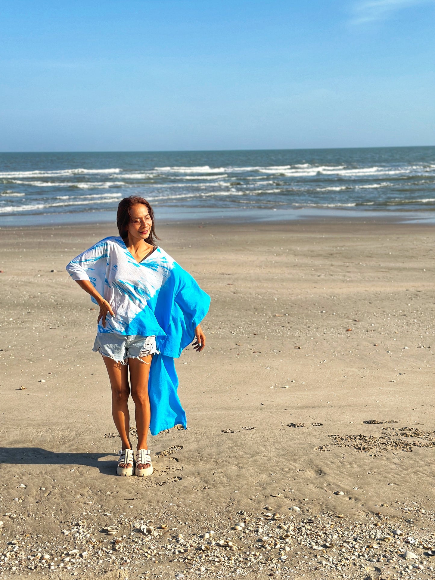 A woman strolls along the beach wearing a light cotton kaftan dress with vibrant blue tie dye, showcasing the short front design.