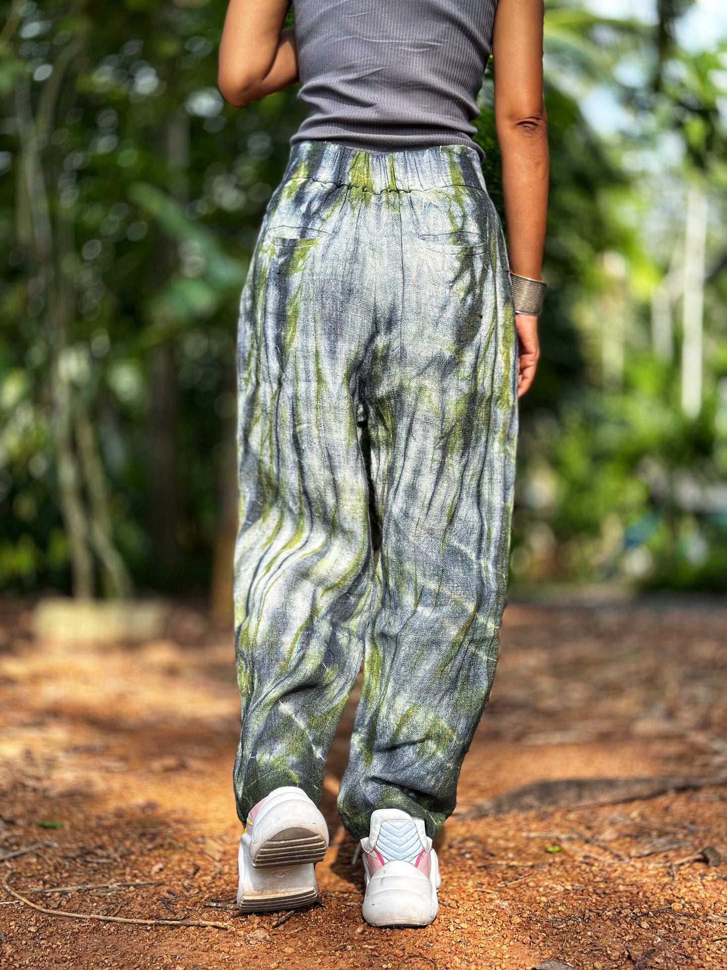 Model in Nuichan tie-dye hemp pants showcasing a gray-green tie-dye pattern on handwoven Hmong hill tribe fabric. These boho hemp trousers, paired with a casual top, create an earthy aesthetic in a lush green outdoor setting.