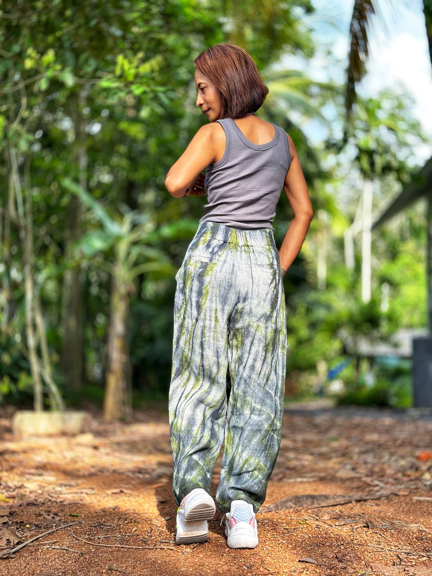 Nuichan tie-dye hemp pants in gray-green, featuring a loose fit with an elastic back and four pockets. These comfortable boho hemp trousers, crafted from handwoven Hmong hill tribe fabric, are styled with a crop top in a serene indoor setting