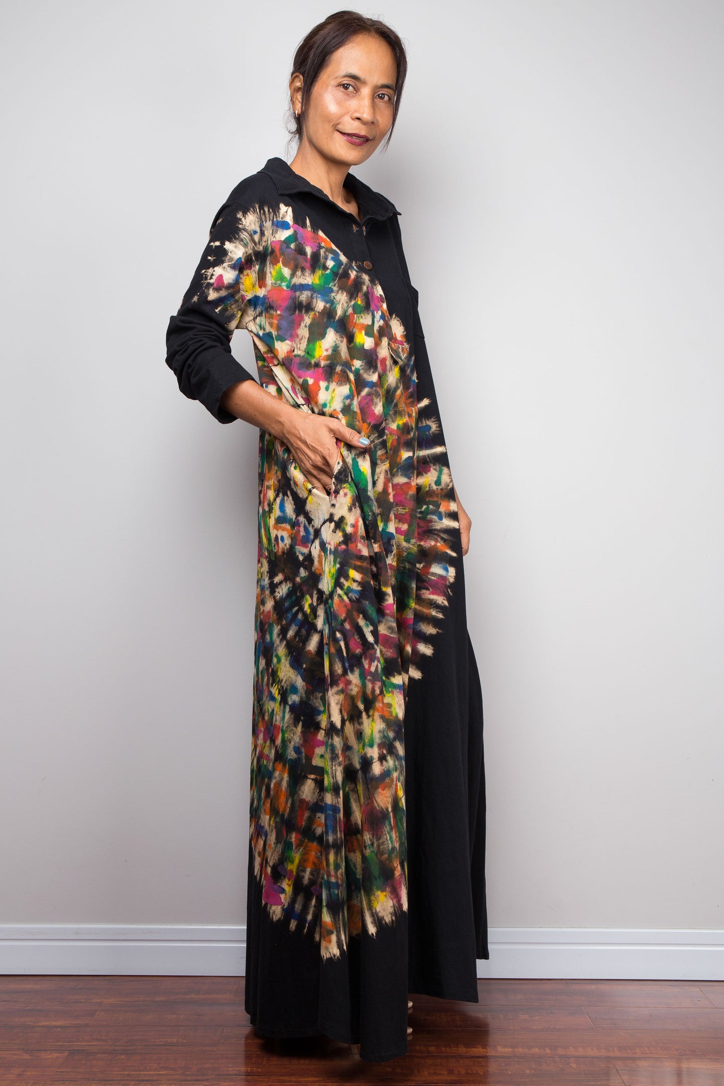 Side front view of woman in a black long-sleeve tie dye maxi shirt dress. An asymmetric, vibrant hand-dyed multi-color tie-dye sunburst sweeps diagonally across the front. She has hands in side pockets.