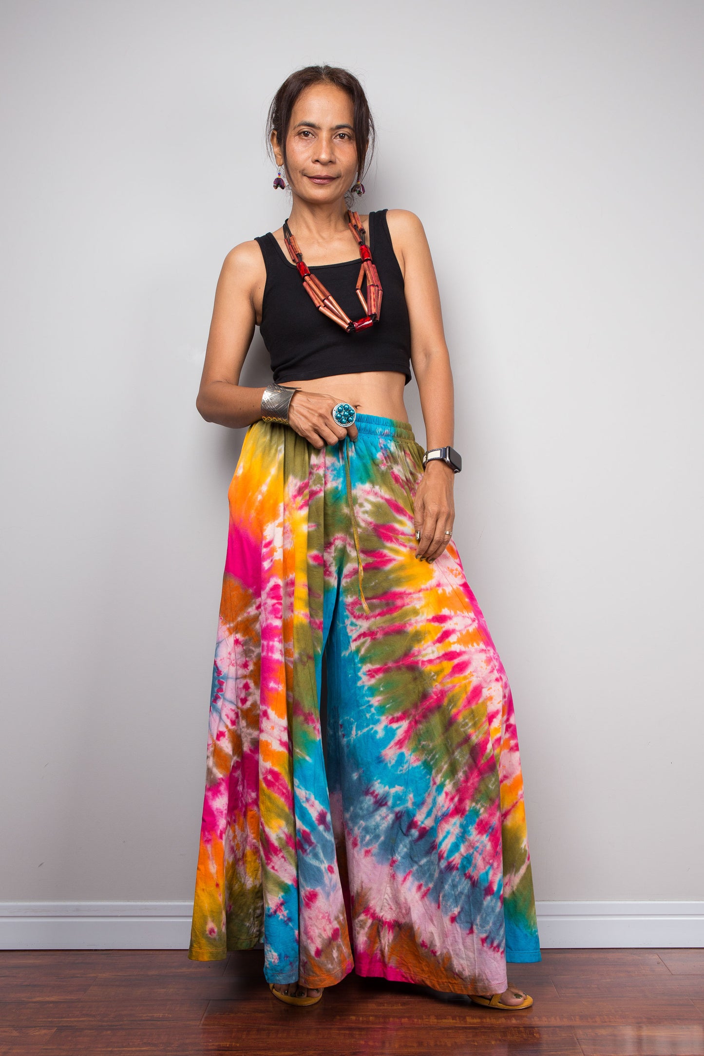 Front view of a petite female model wearing the Nuichan rainbow carnival tie dye wide leg palazzo pants, showcasing the complex multi-point starburst hand-dyed pattern in hot pink magenta turquoise orange and olive, dramatic floor-length ultra-wide leg silhouette, and distinctive dual-tone smocked elastic waistband in turquoise and olive, styled with a black crop top