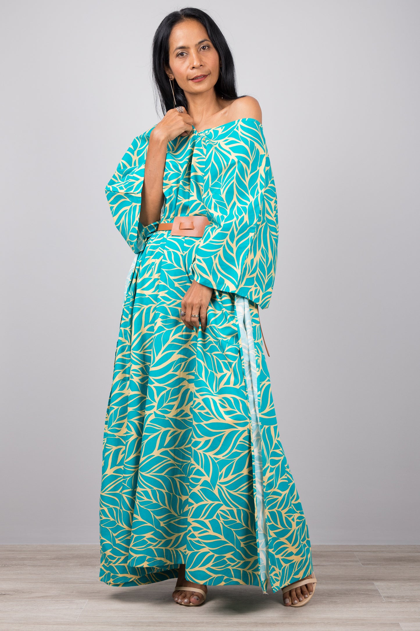 Woman in a versatile turquoise kaftan dress with yellow leaf pattern, standing against a gray wall with a wooden floor, accessorized with a smartwatch and bracelet.