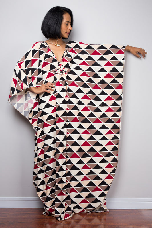 A woman wearing a black, red, beige, and brown geometric triangle-patterned kaftan dress, standing against a plain gray wall on a wooden floor, with wide, flowing sleeves and a V-neckline, accessorized with a gold necklace and earrings.