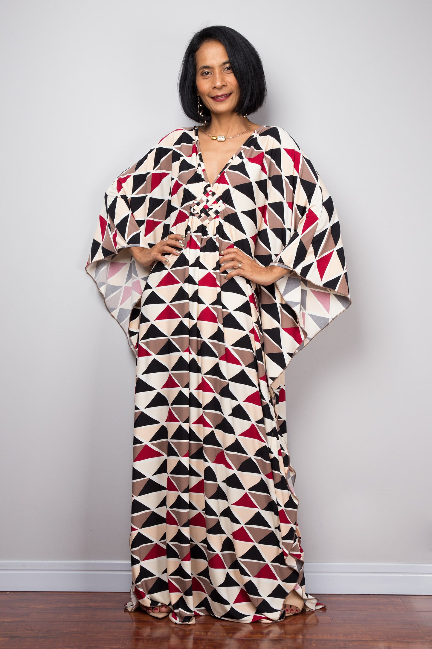 Woman in a black, red, and beige geometric triangle kaftan dress by Nuichan, standing with hands on hips, showcasing the dress’s wide sleeves and maxi length, in a studio with a gray backdrop and wooden floor.