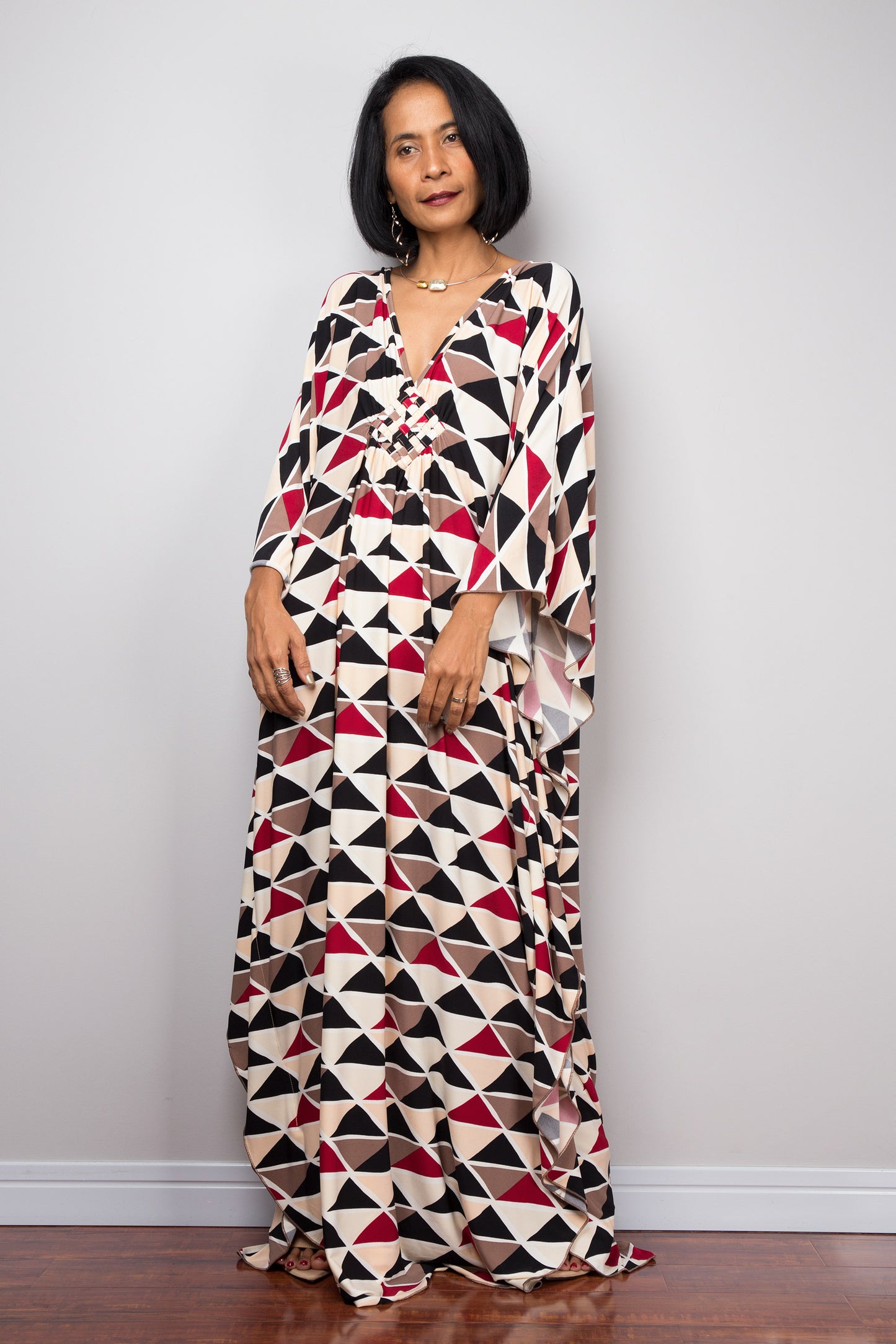 Nuichan’s black, red, and beige triangle-patterned kaftan dress, modeled by a woman with short black hair, posed gracefully in a minimalist studio with a gray background and dark wood flooring.