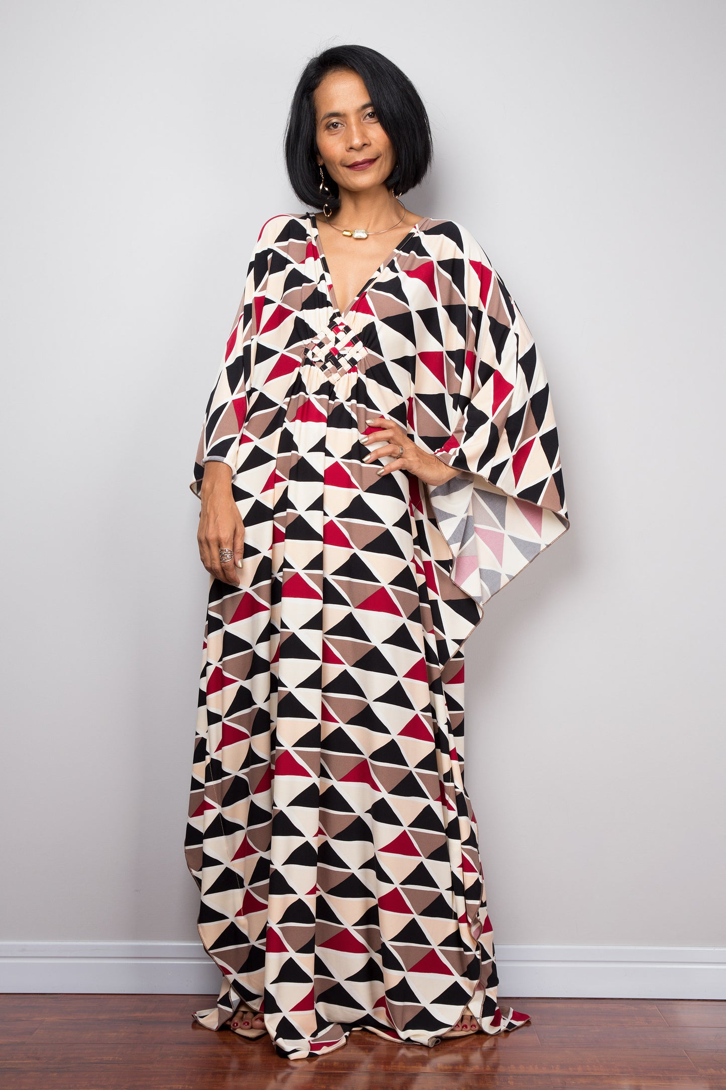 Nuichan’s black, red, and beige triangle-patterned kaftan dress, worn by a woman with dark hair and gold jewelry, posed against a gray wall on a dark wood floor, highlighting its bold boho style.