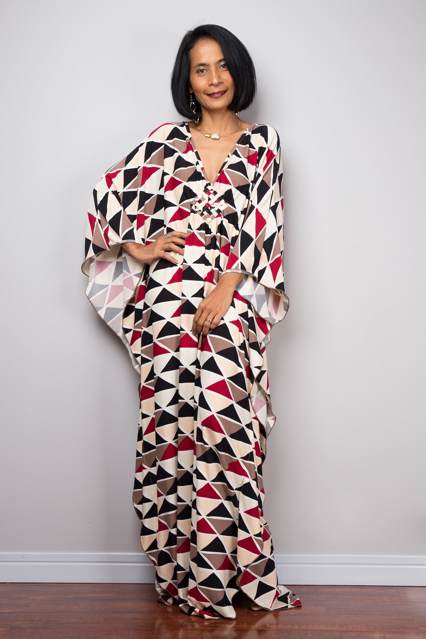 A woman modeling a black, red, beige, and brown triangle-patterned kaftan dress from Nuichan, with a loose fit, V-neck, and flowing sleeves, standing in a simple gray and wood studio setting, accessorized with a necklace.