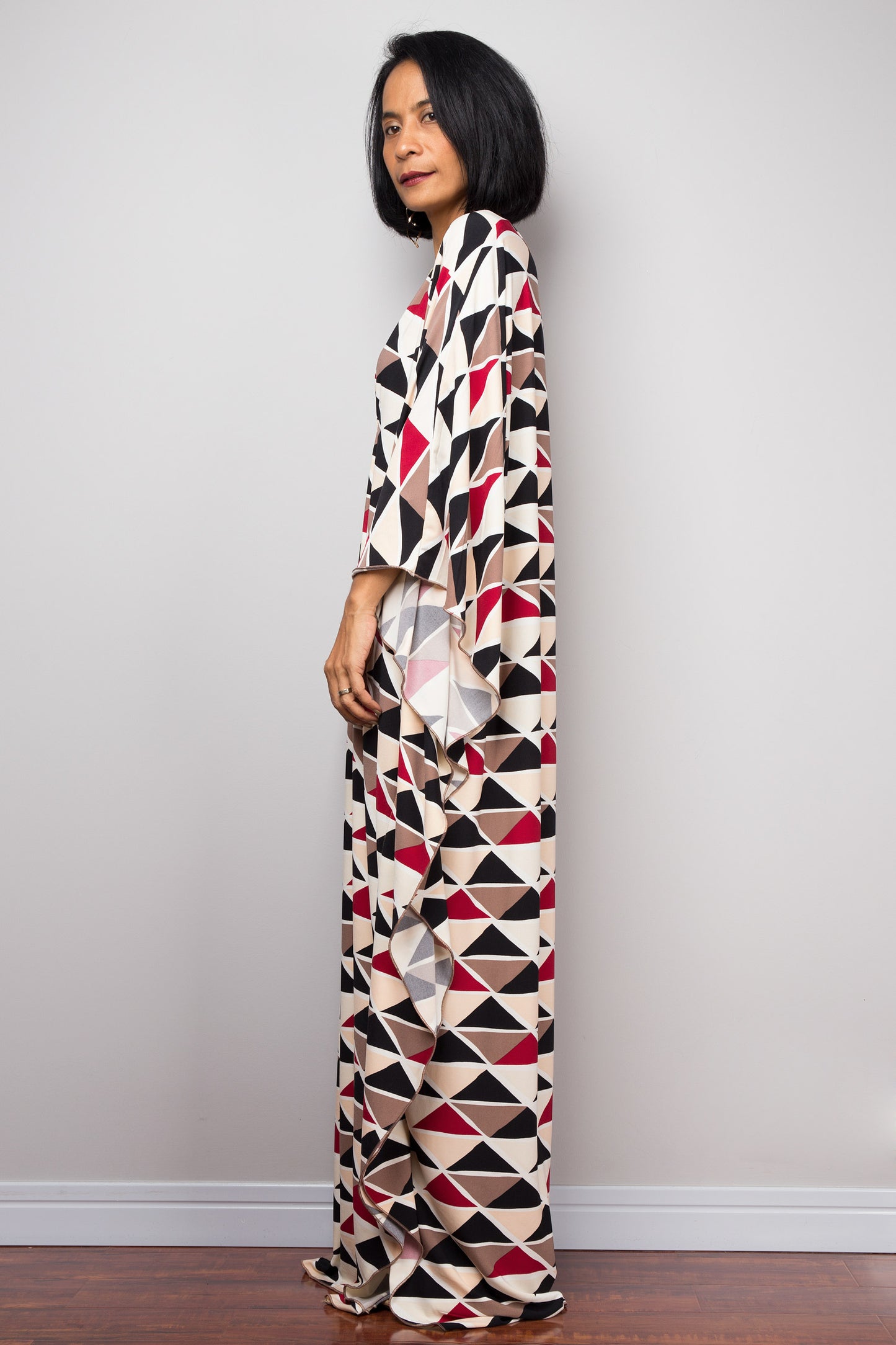 A floor-length black, red, beige, and brown triangle-patterned kaftan dress from Nuichan, worn by a woman standing sideways, featuring loose, flowing fabric and short sleeves, set against a gray wall and wooden floor.