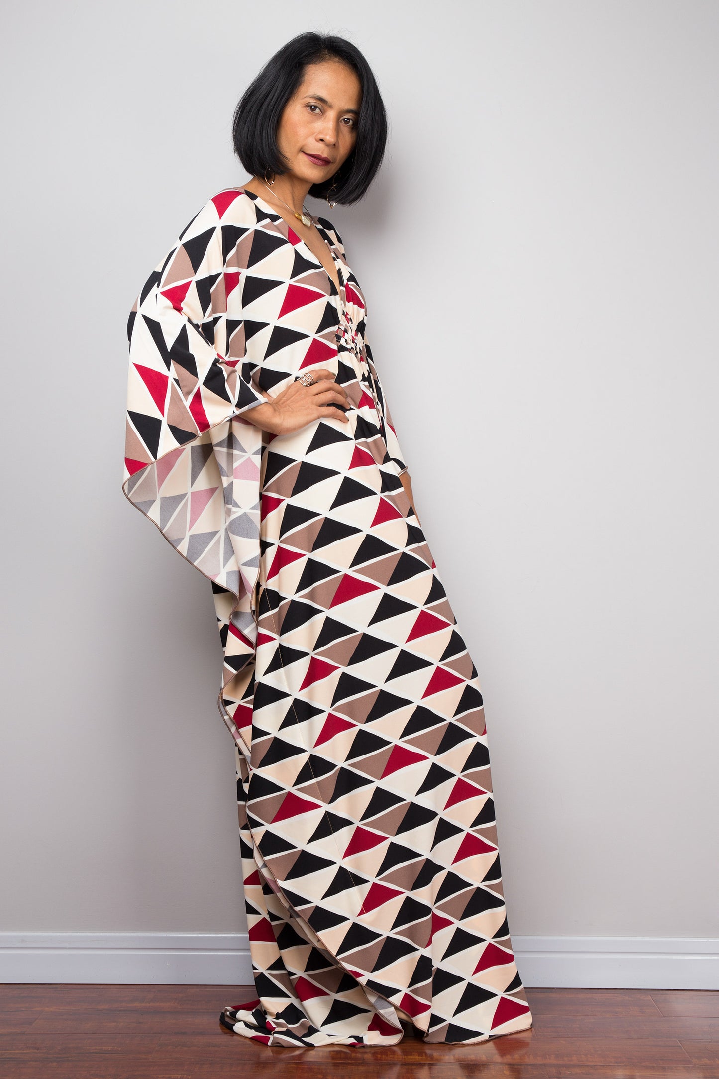 Black, red, beige, and brown triangle-patterned kaftan dress by Nuichan, worn by a woman with short black hair, standing gracefully against a gray wall and wooden floor, featuring wide sleeves and a floor-length design.