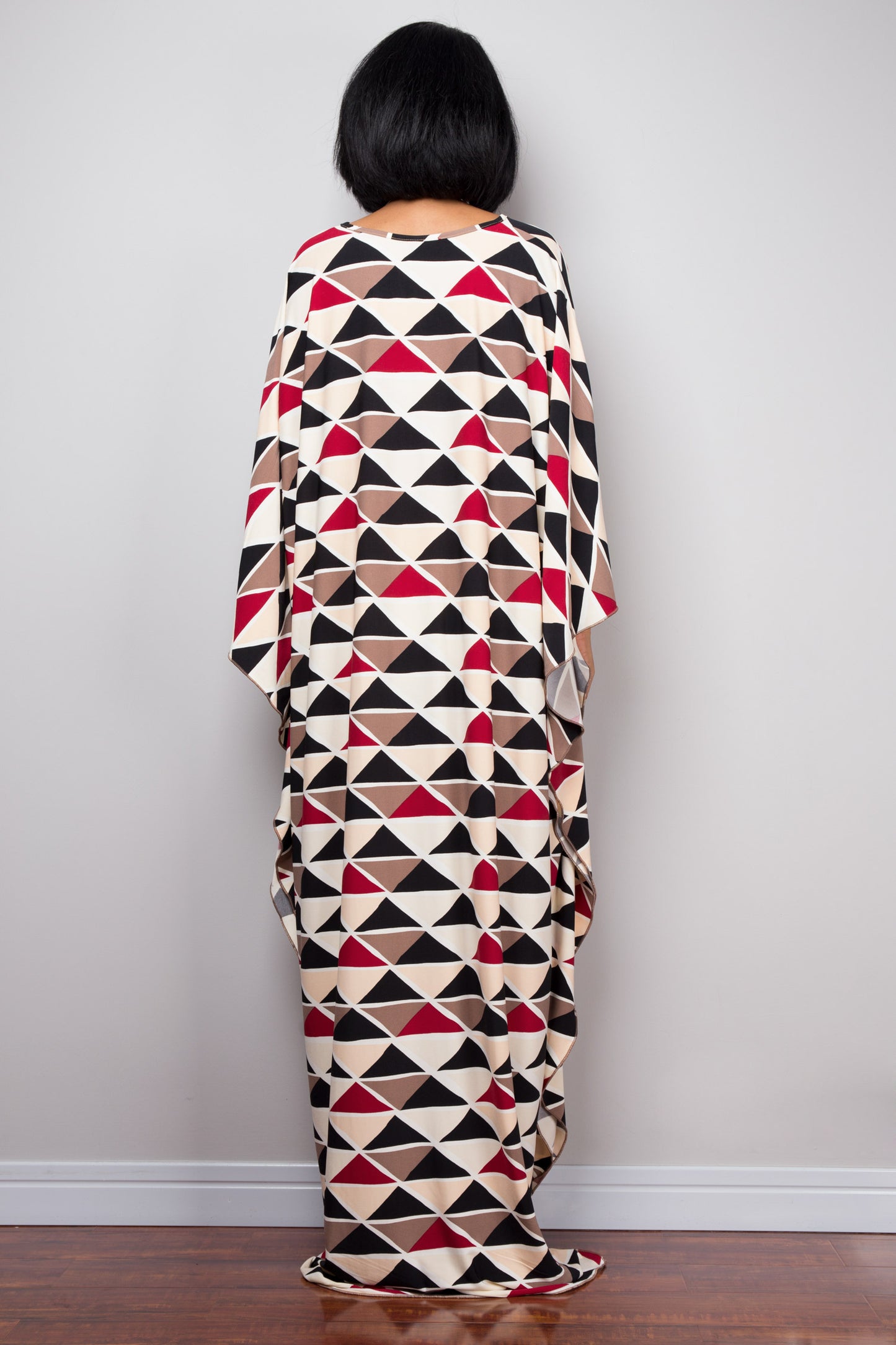 Black, red, beige, and brown triangle-patterned kaftan dress from Nuichan, worn by a woman with dark hair and gold jewelry, back view pose against a gray studio wall and dark wood floor, highlighting its vibrant boho-chic design and flowing silhouette.
