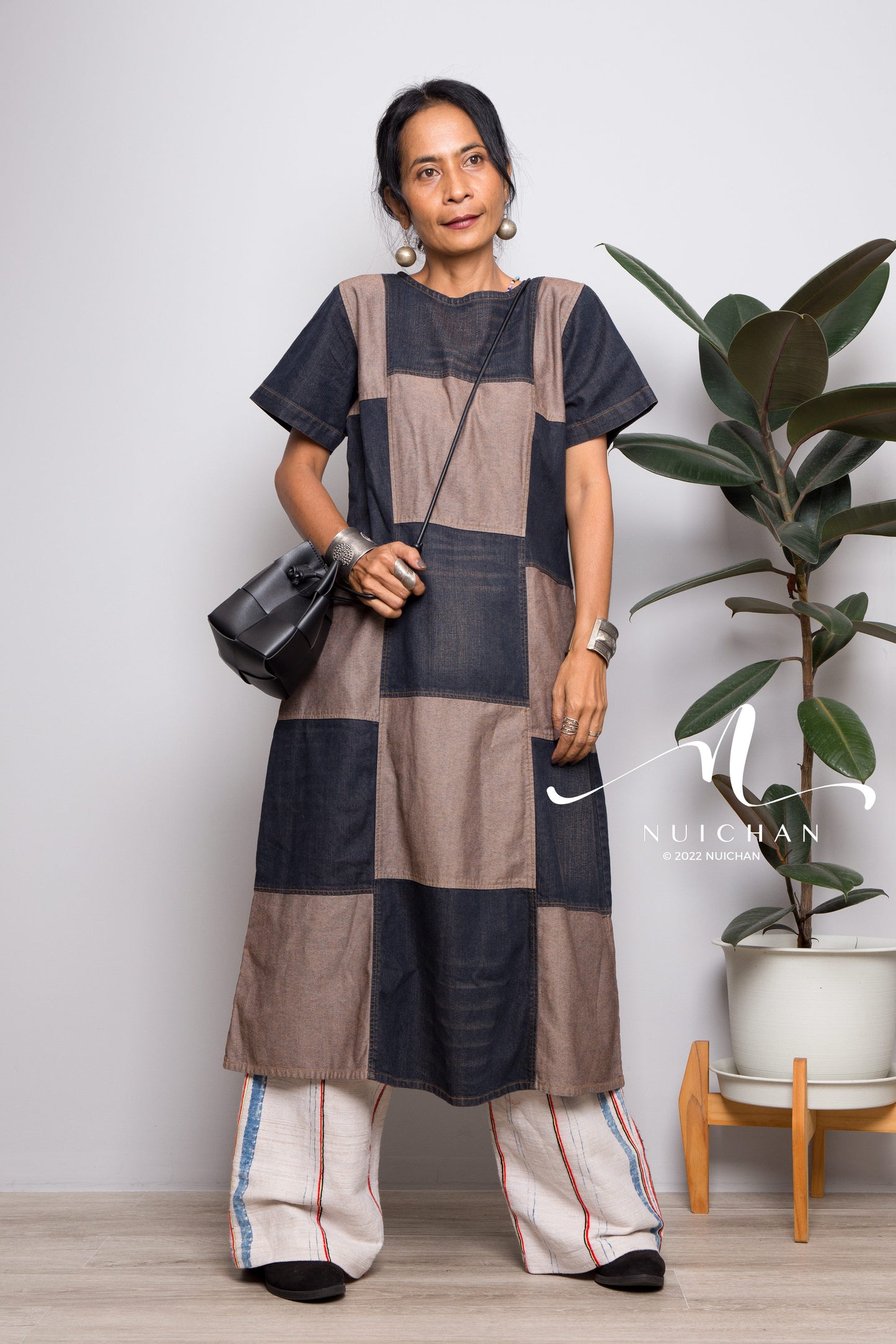 A denim patchwork mid-length dress by Nuichan, featuring a stonewashed short-sleeve design in two-tone blue. This boho dress, set against a forest backdrop with a stream, exudes a free-spirited, earthy vibe.