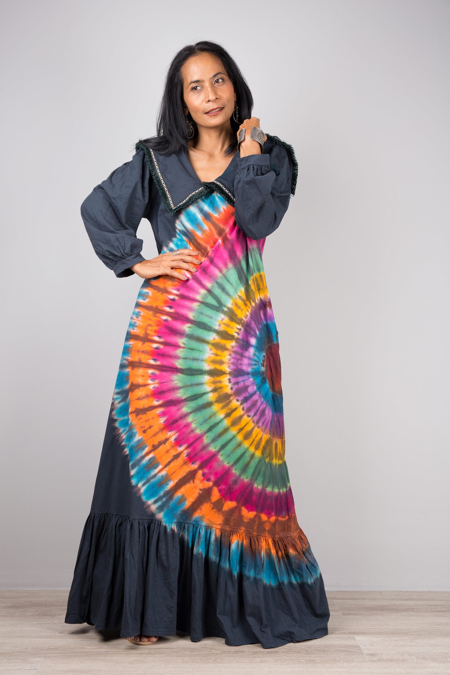 Nuichan rainbow tie-dye kaftan maxi dress with a loose fit, navy-trimmed collar, and long sleeves, modeled by a woman against a gray wall and wooden floor, paired with nude sandals.