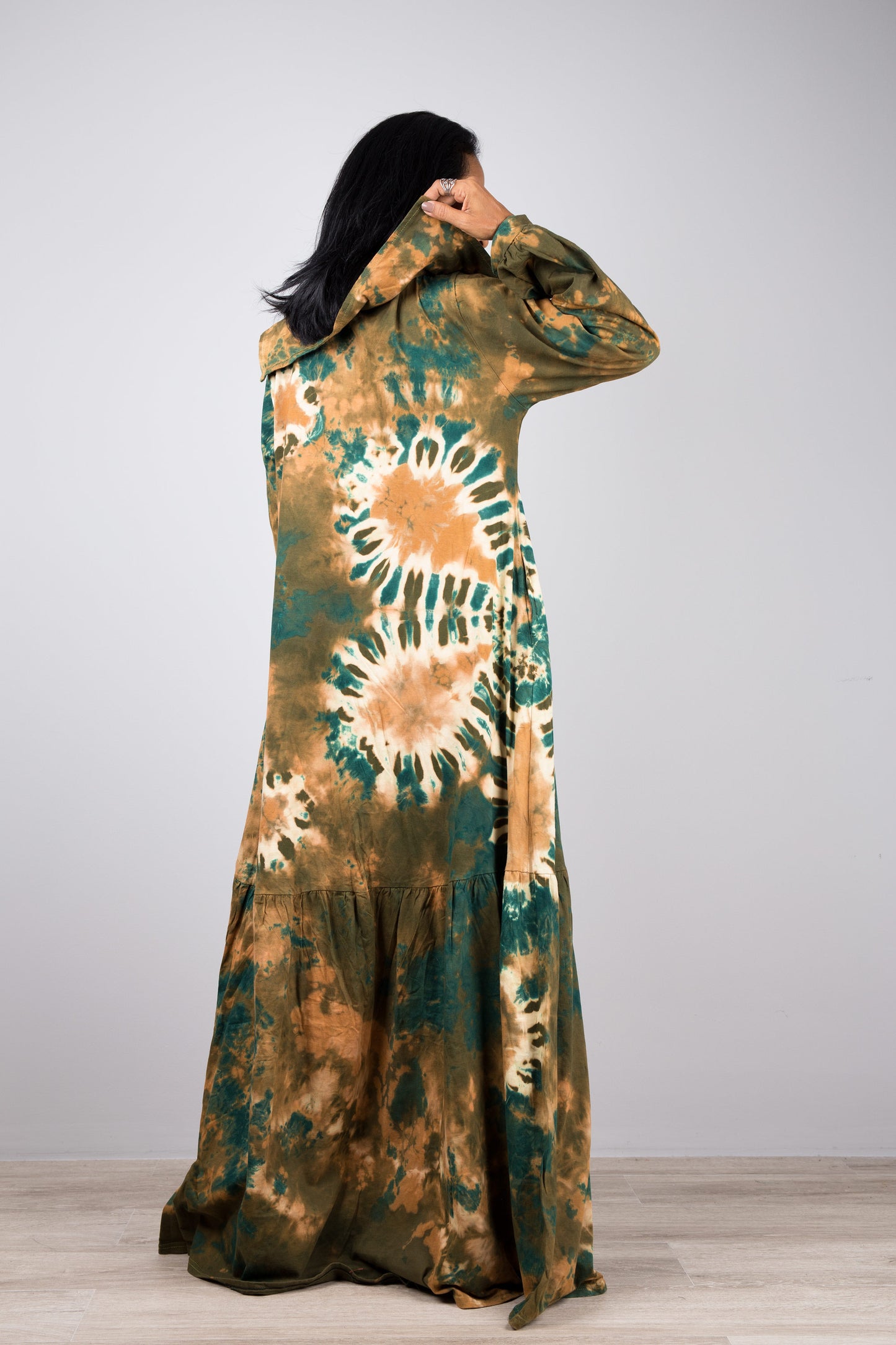 Green tie-dye kaftan maxi dress with a navy-trimmed V-neck and long sleeves, worn by a woman with nude sandals and earrings, set against a minimalist gray and wood interior.