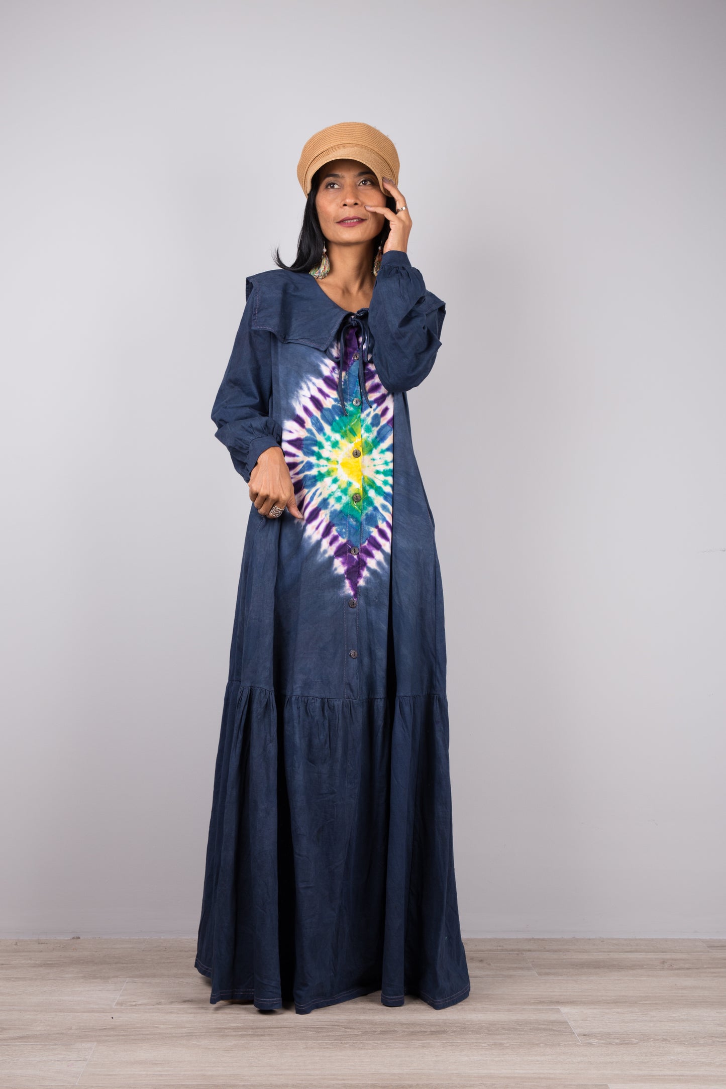 Dark blue tie-dye kaftan maxi dress with a navy-trimmed V-neck and long sleeves, worn by a woman with nude sandals and earrings, set against a minimalist gray and wood interior